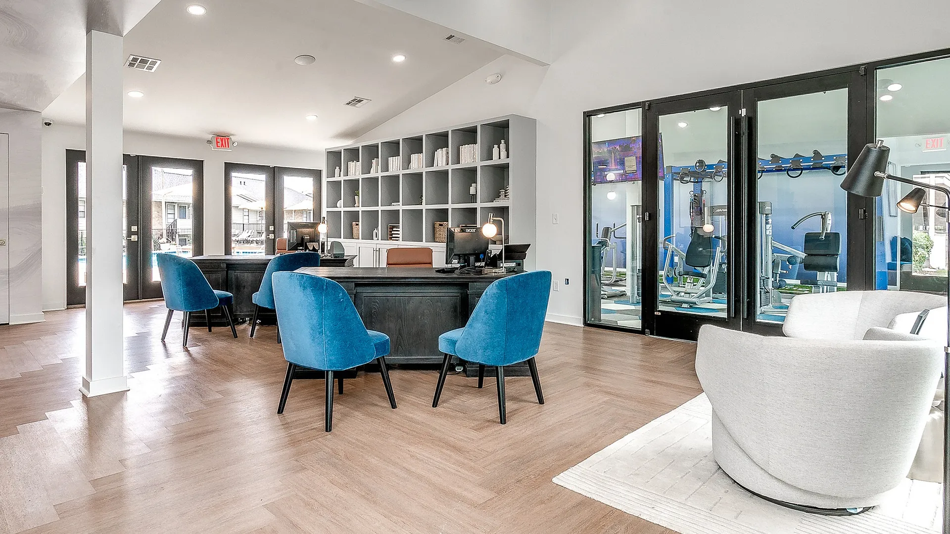 Elegant communal area featuring modern seating with vibrant blue chairs, expansive windows, and a sleek design overlooking a fitness center.