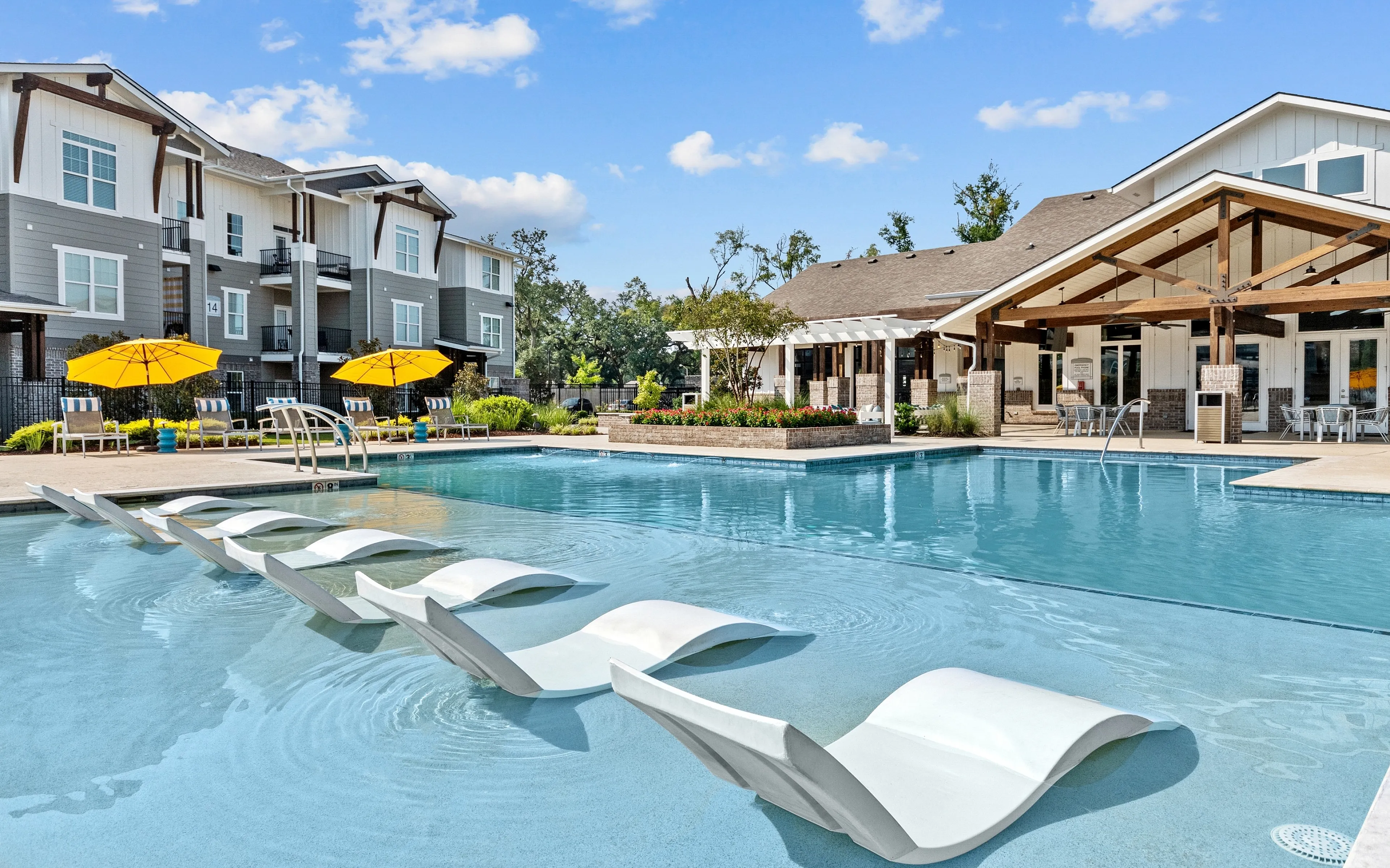 Luxury apartment complex with a sparkling swimming pool, modern lounge chairs, vibrant yellow umbrellas, and elegant architecture under a clear blue sky.