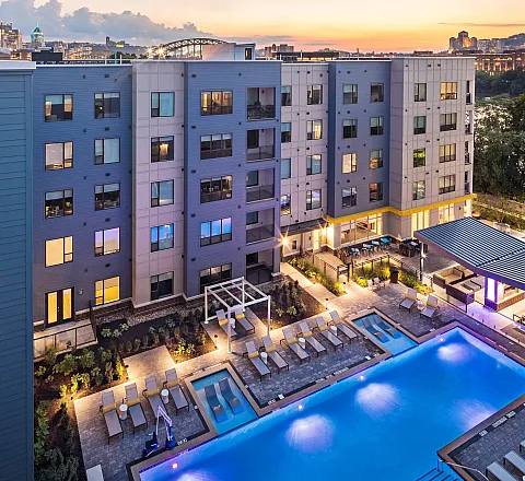 Modern apartment complex with a spacious swimming pool, stylish lounge areas, and cityscape views at sunset.