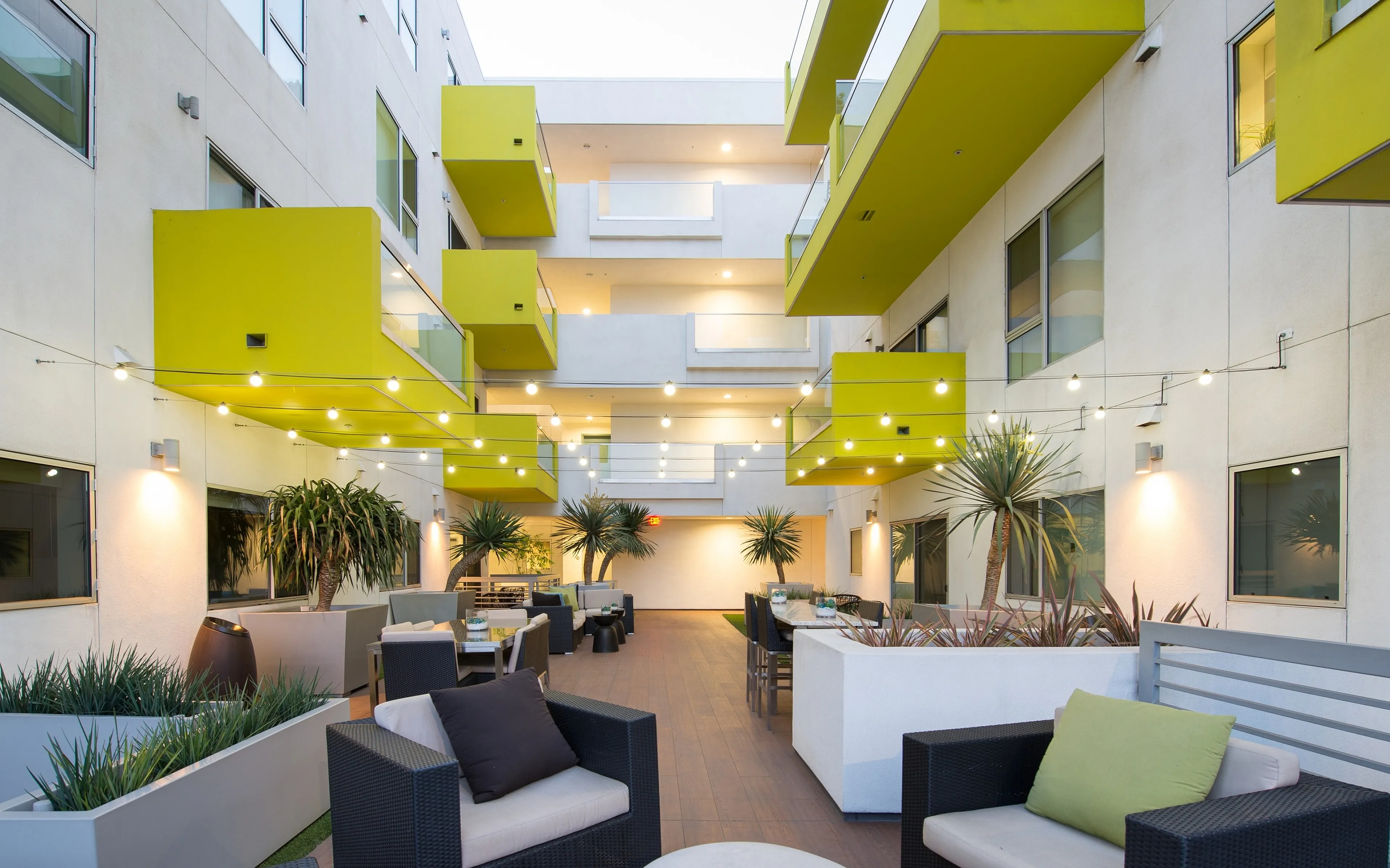 Modern apartment courtyard with stylish seating, vibrant greenery, and contemporary yellow balconies, beautifully lit by overhead string lights.