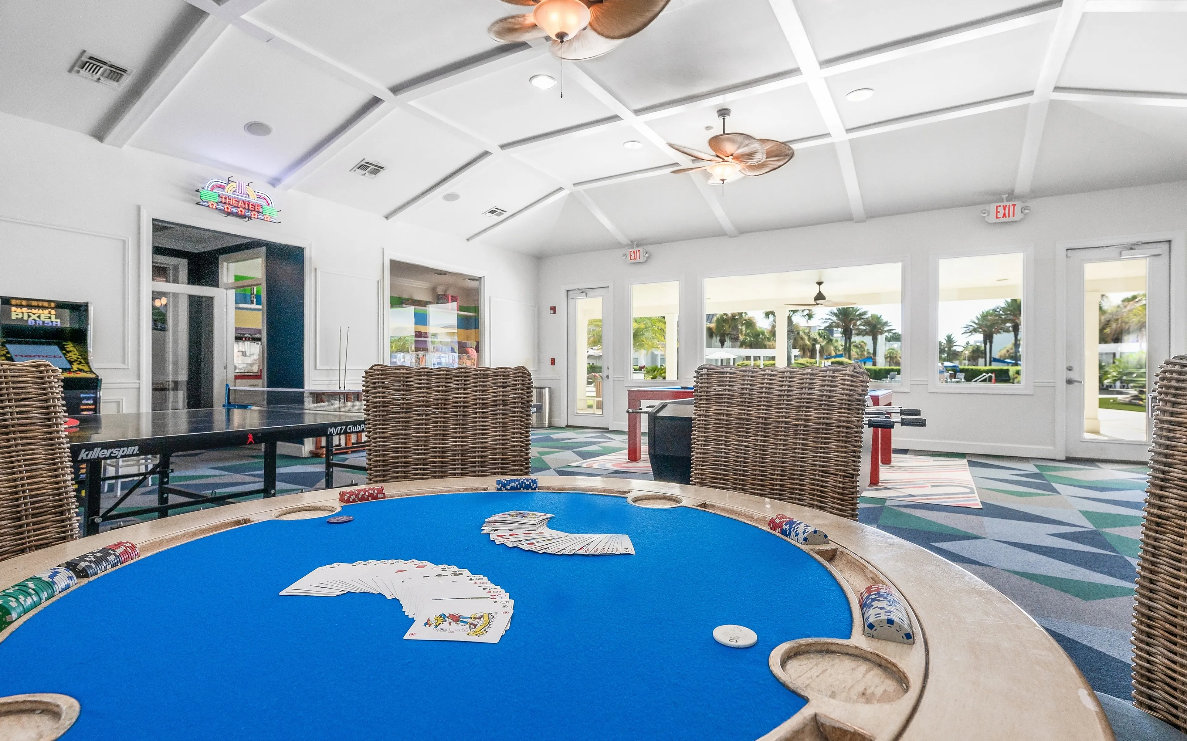 Vibrant game room featuring a poker table with cards and chips, ping pong table, arcade games, and large windows with a scenic view.