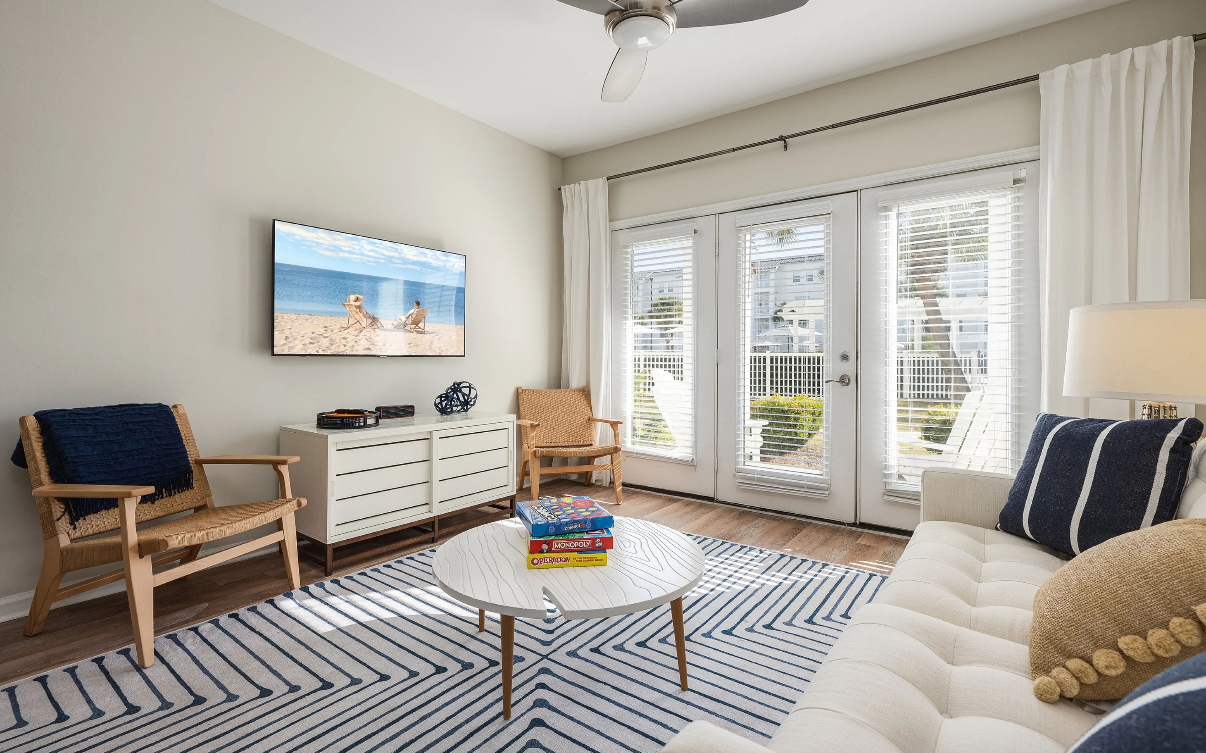 Bright and inviting living room with a cozy cream sofa, stylish wooden chairs, and a chic striped rug. Sunlit space features a wall-mounted TV, modern ceiling fan, and large windows with a view of a vibrant outdoor area. Perfect for relaxat