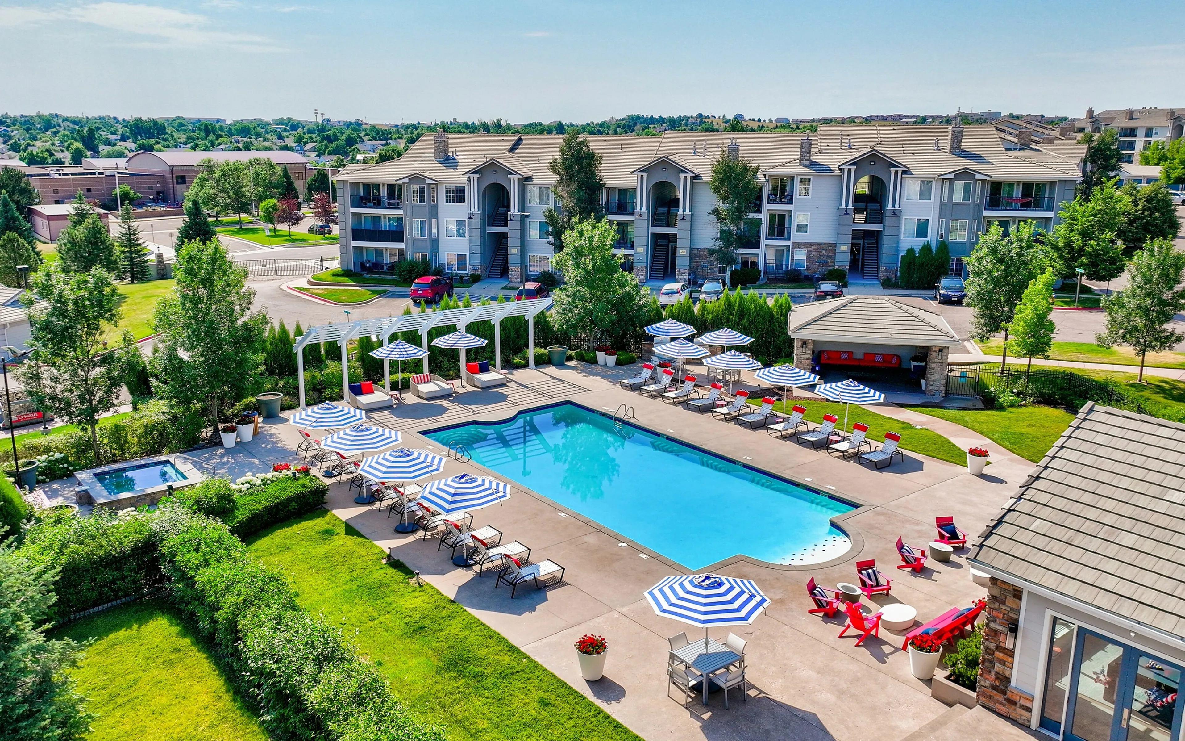 Scenic apartment complex featuring a sparkling swimming pool, stylish lounge area with umbrellas, lush greenery, and modern buildings in the background.