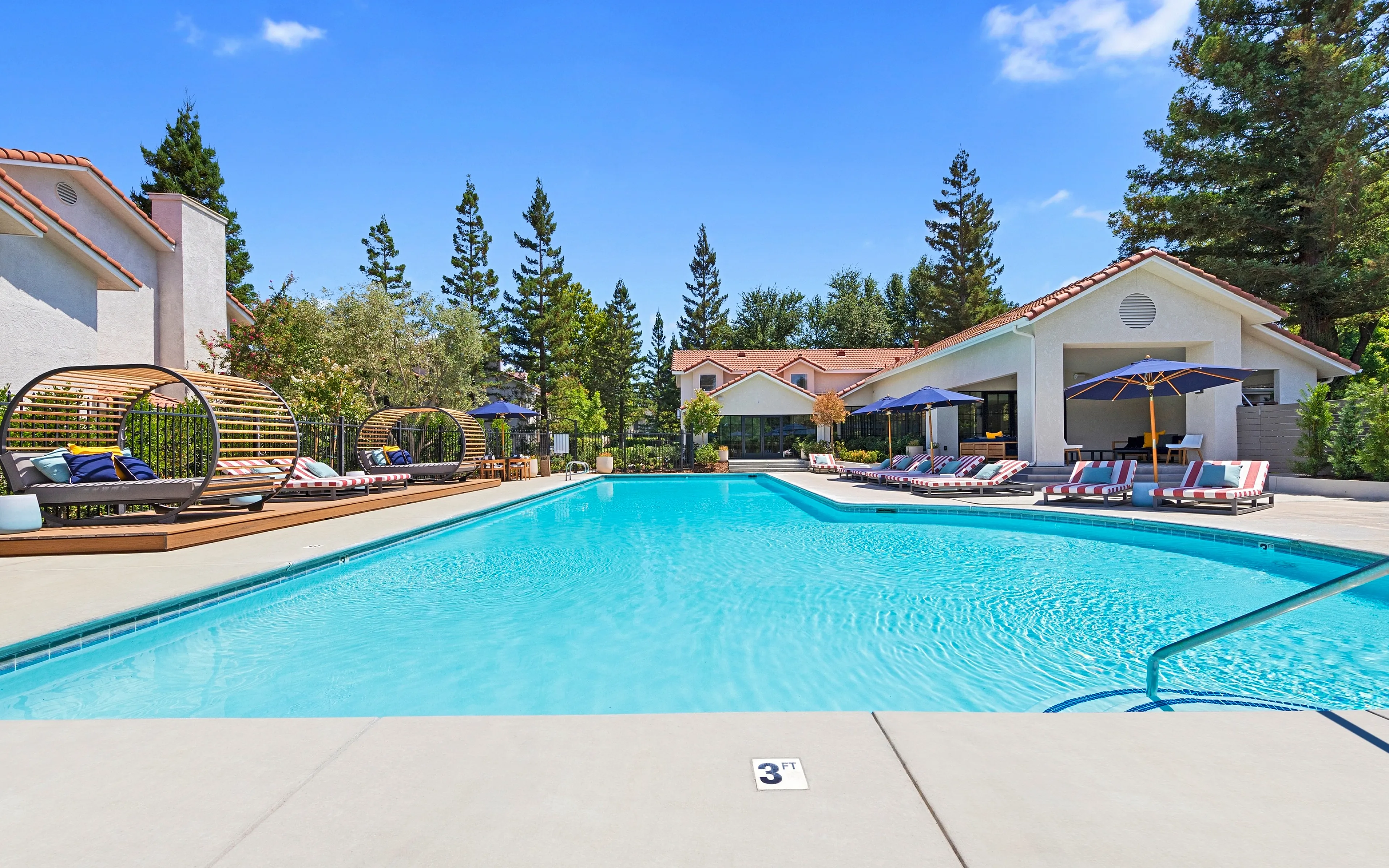 Luxurious outdoor swimming pool surrounded by stylish sun loungers and modern cabanas, nestled amidst serene pine trees and charming villas under a bright blue sky.