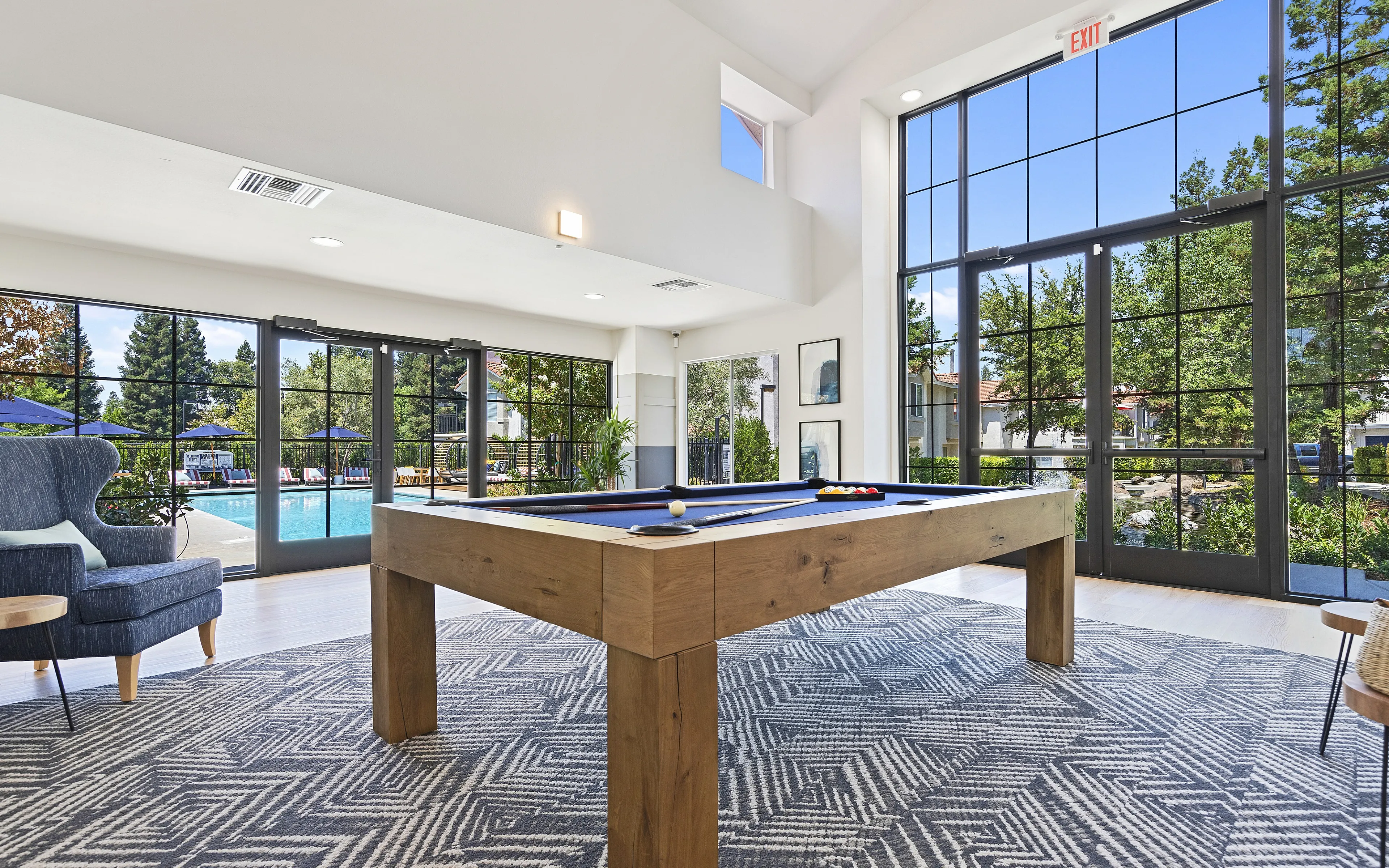 Modern game room with stylish pool table, elegant armchair, and expansive glass windows overlooking a sparkling outdoor pool.