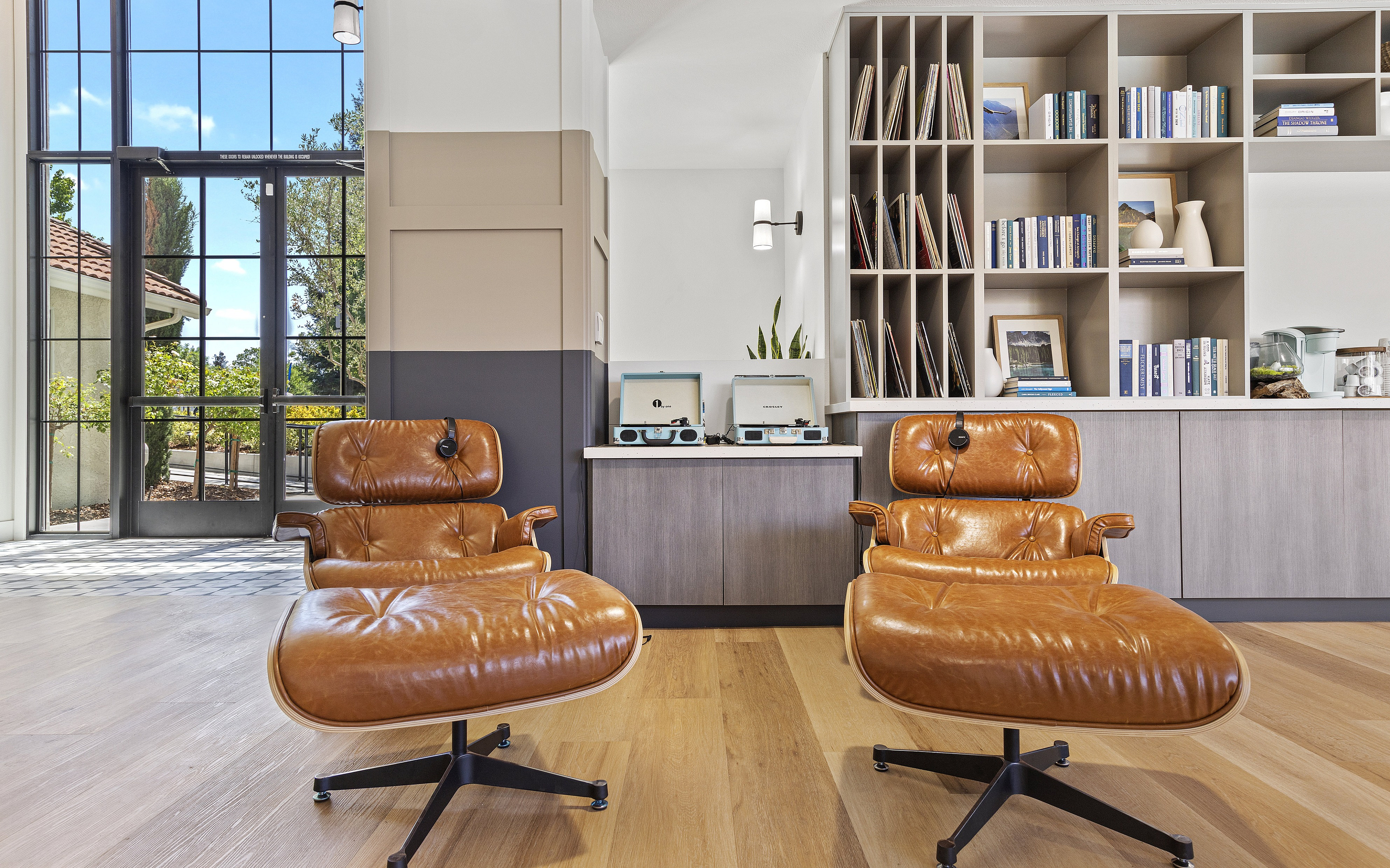 Contemporary lounge area with elegant leather lounge chairs, sleek shelving with books and decor, large sunlit windows, and a stylish wood floor.
