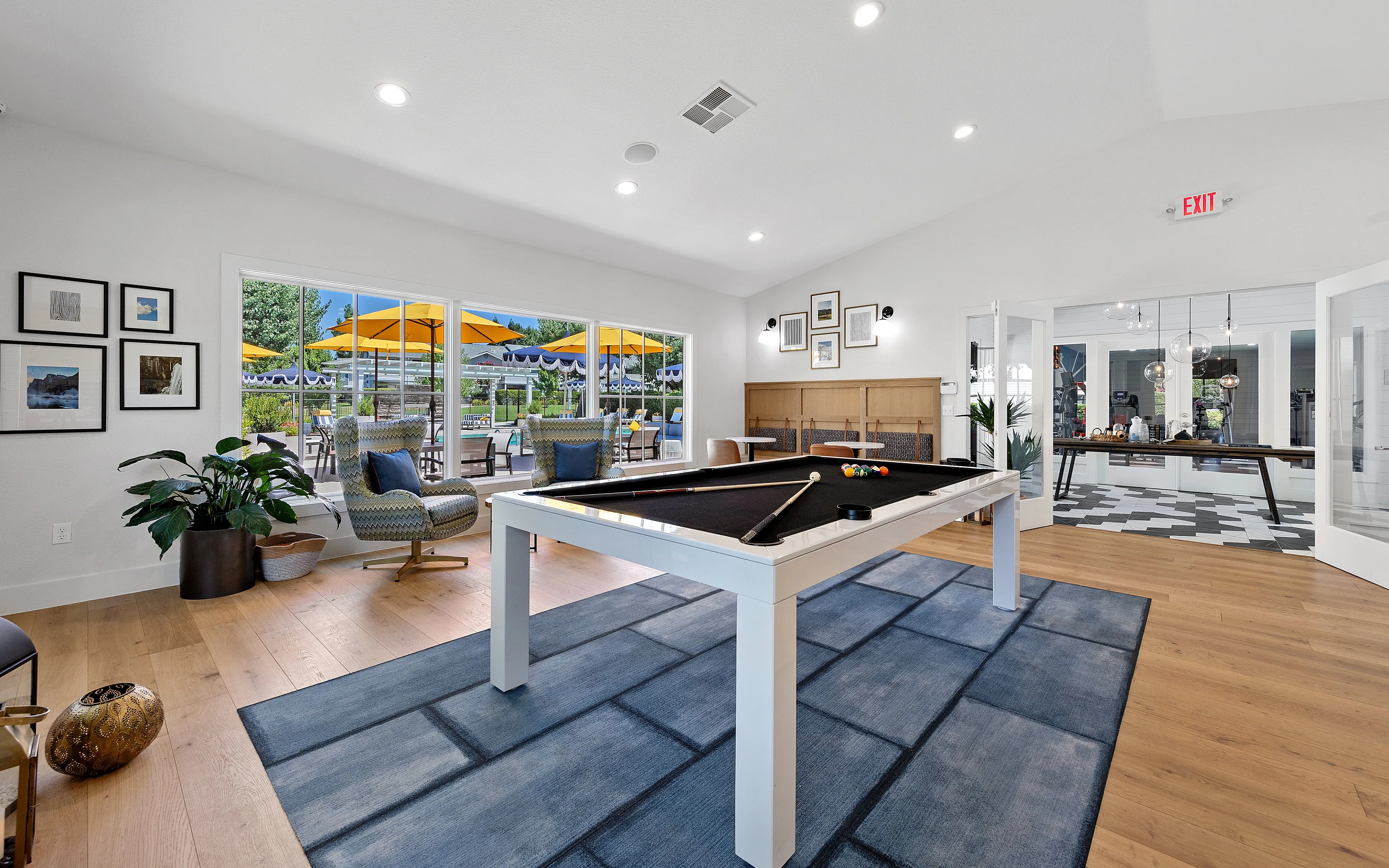 Chic recreation area with a modern pool table, bright seating, and large windows overlooking a vibrant outdoor patio with yellow umbrellas.