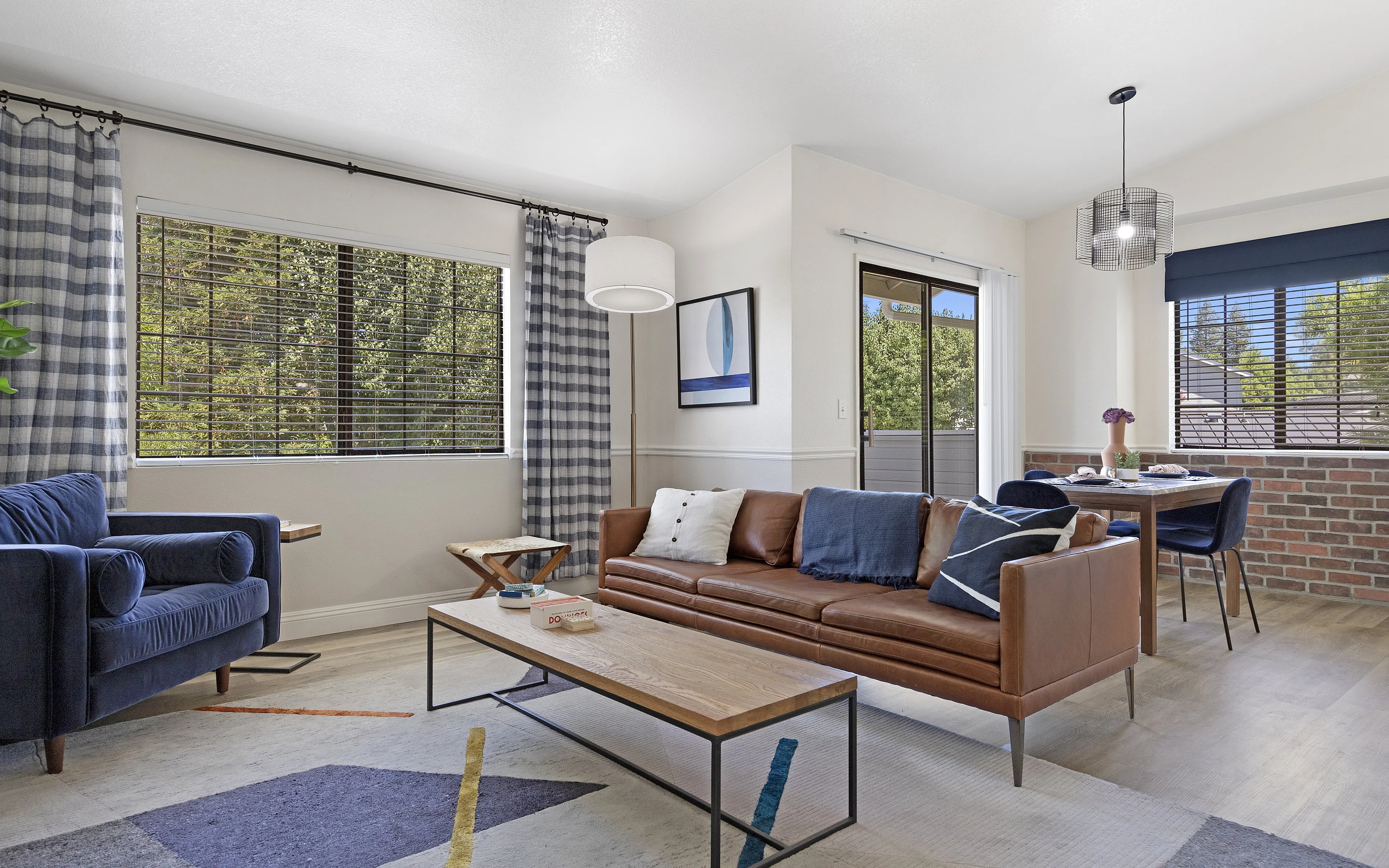 Bright and inviting living space featuring a stylish leather sofa, plush blue armchair, and a modern dining area with large windows showcasing a lush green view.
