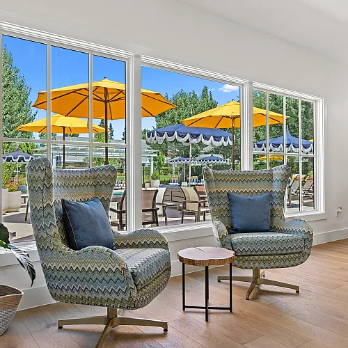 Charming living area with cozy armchairs and stylish decor, overlooking a vibrant courtyard through expansive windows, featuring bright yellow umbrellas and lush greenery.
