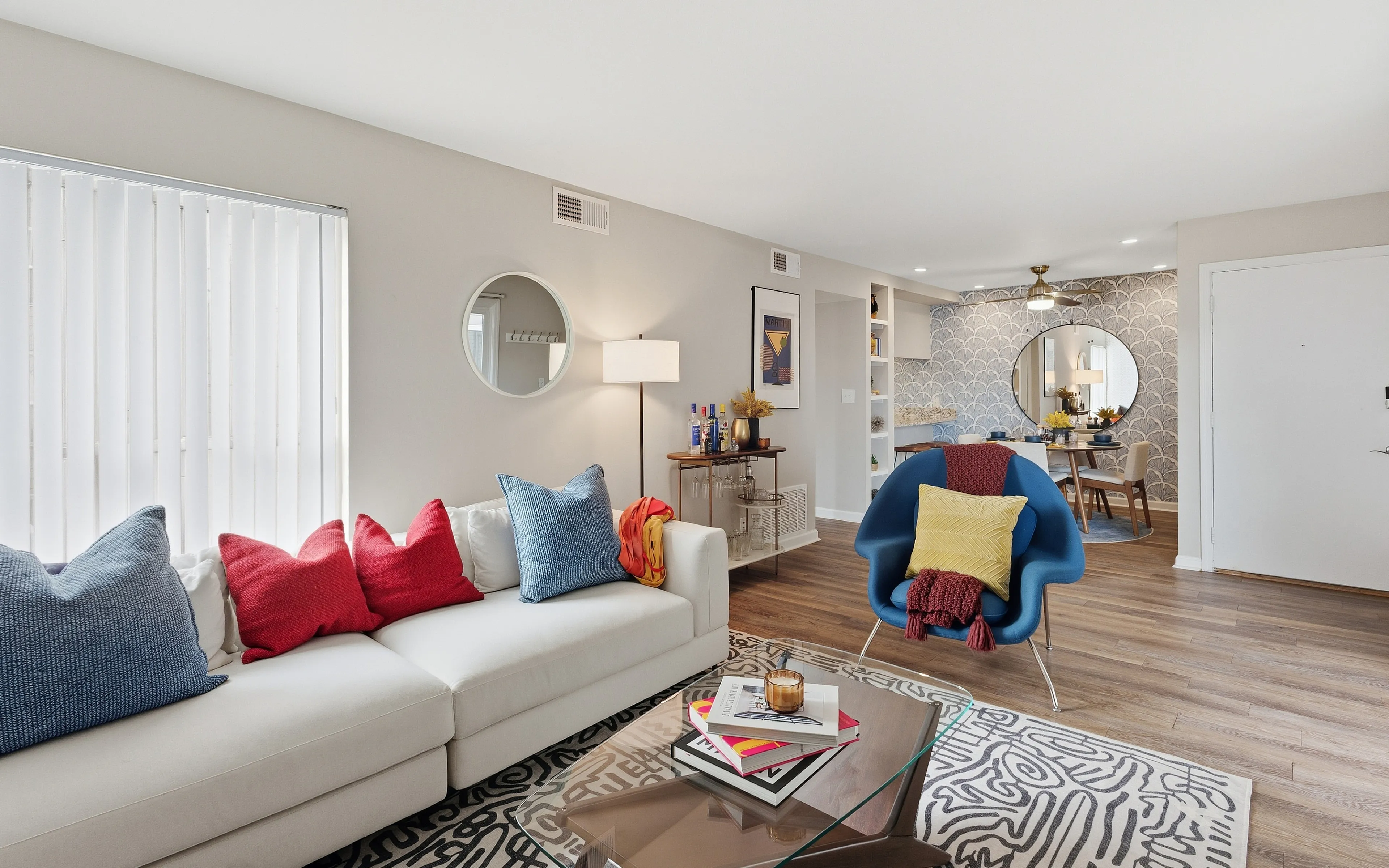 Modern living room with vibrant decor, featuring a cozy white sofa adorned with colorful cushions, a sleek glass coffee table, and a chic blue armchair, leading into a stylish dining area with elegant lighting and a decorative mirrored wall