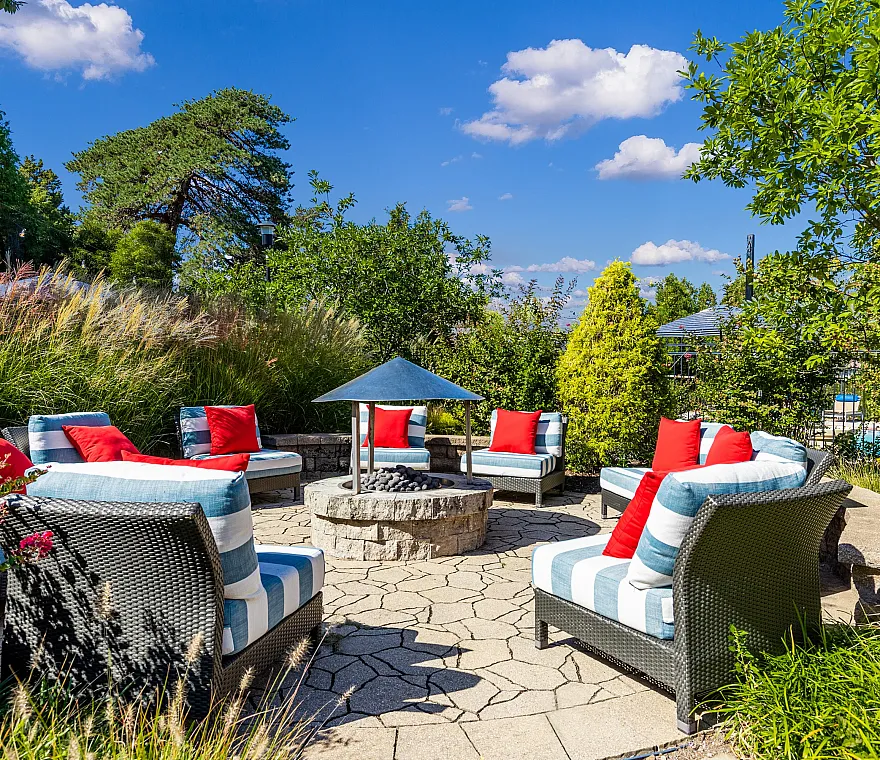 Charming garden patio with stylish wicker seating, vibrant red cushions, and a central fire pit, surrounded by lush greenery under a bright blue sky.