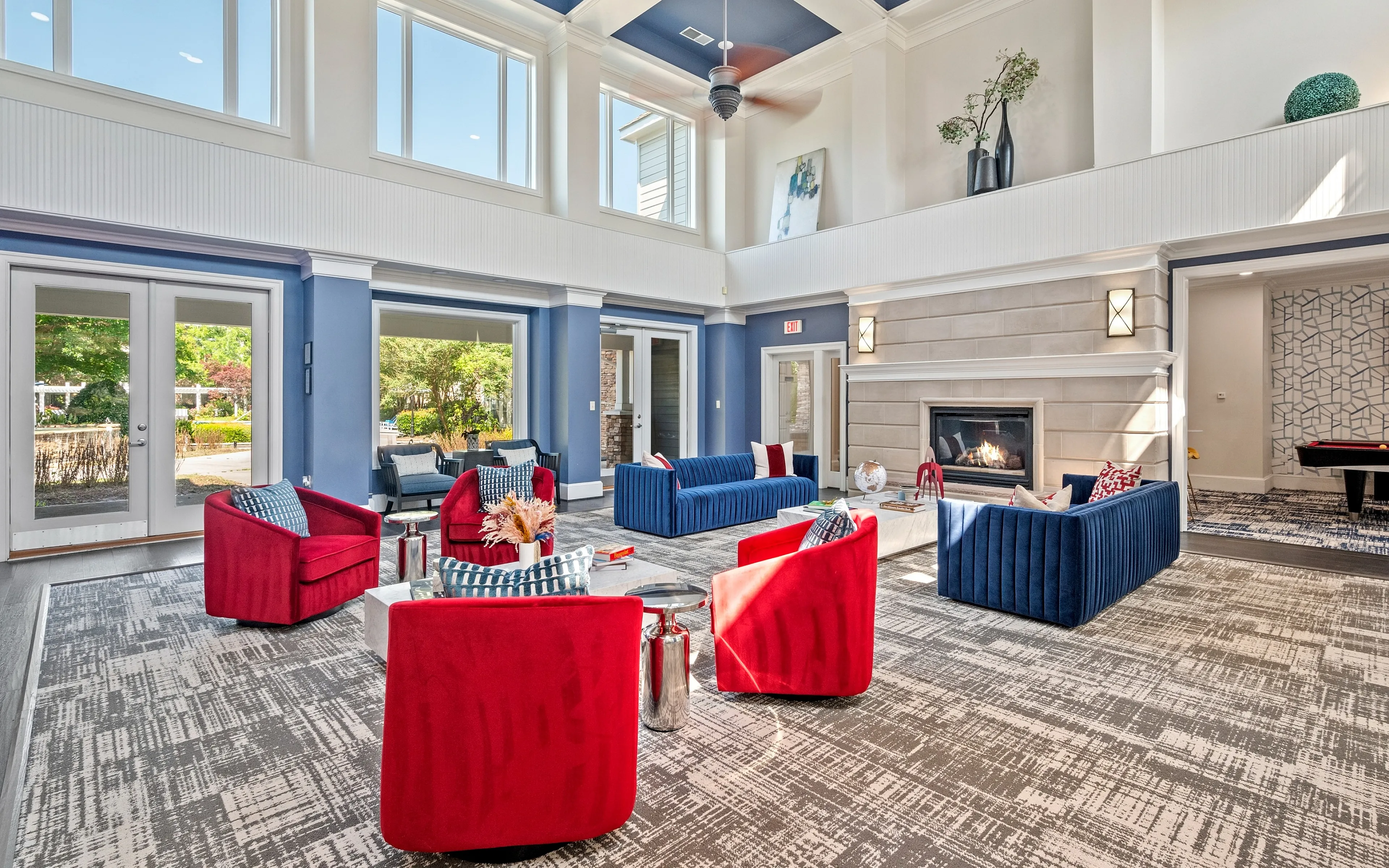 Elegant apartment lounge featuring red and blue seating, high ceilings, large windows, and a cozy fireplace.