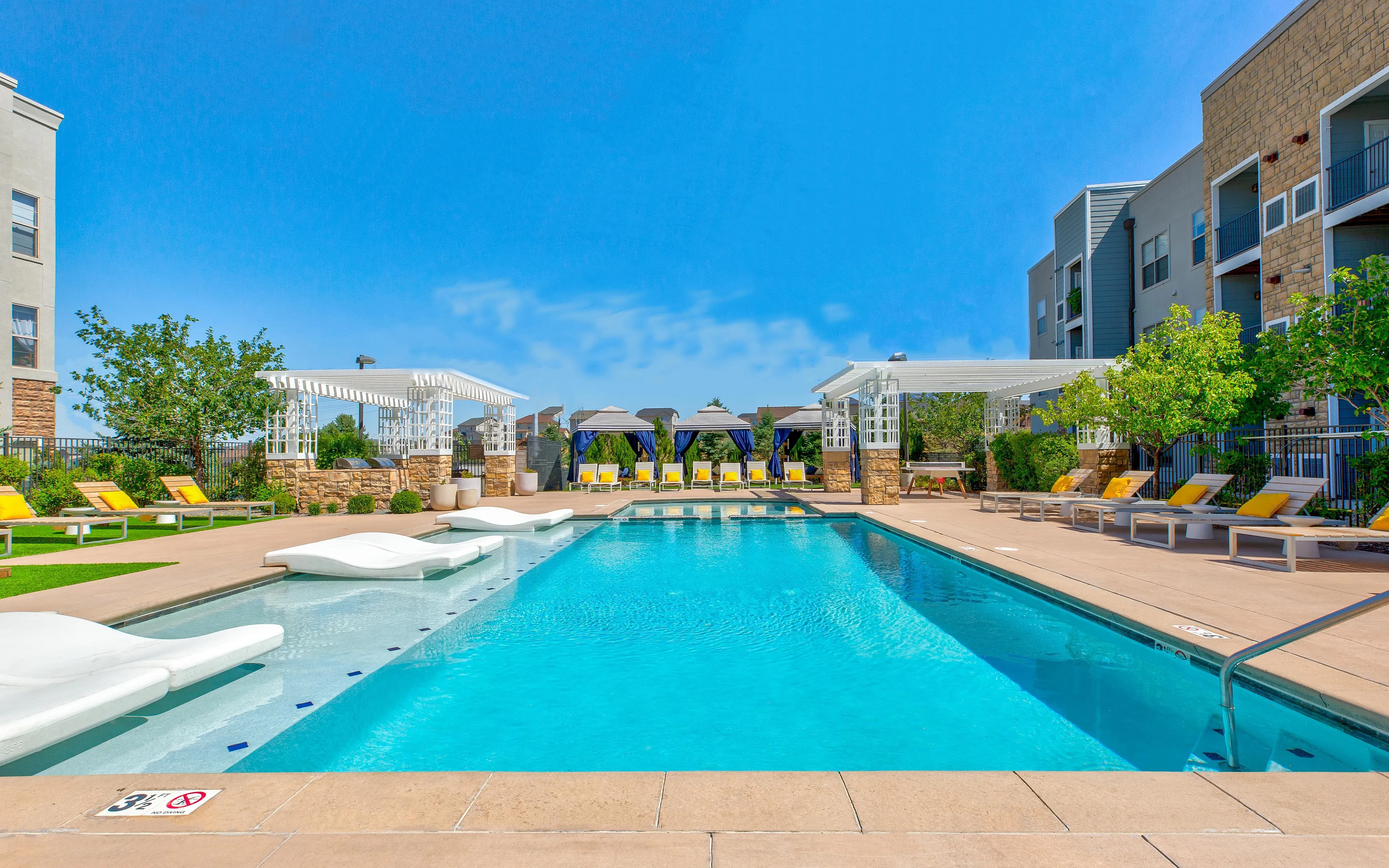 Luxurious apartment complex featuring a large sparkling pool with stylish lounge chairs and elegant cabanas under a clear blue sky.