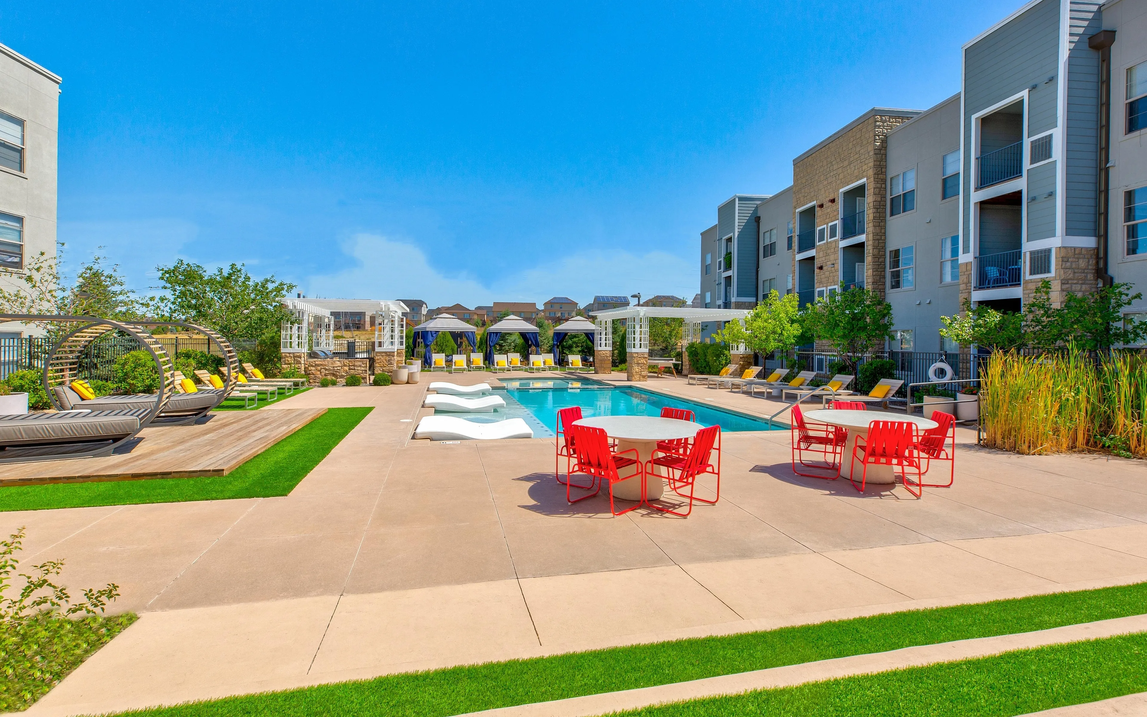 Vibrant apartment complex featuring a sparkling pool, modern lounge chairs, stylish seating, and lush landscaping under a clear blue sky.