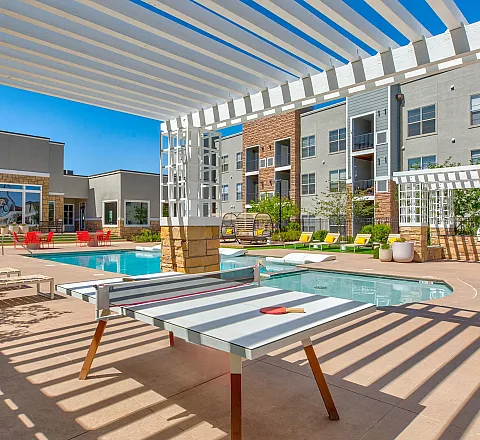 Modern apartment complex featuring a luxurious outdoor pool area, stylish lounge furniture, and a shaded ping pong table under a bright blue sky.
