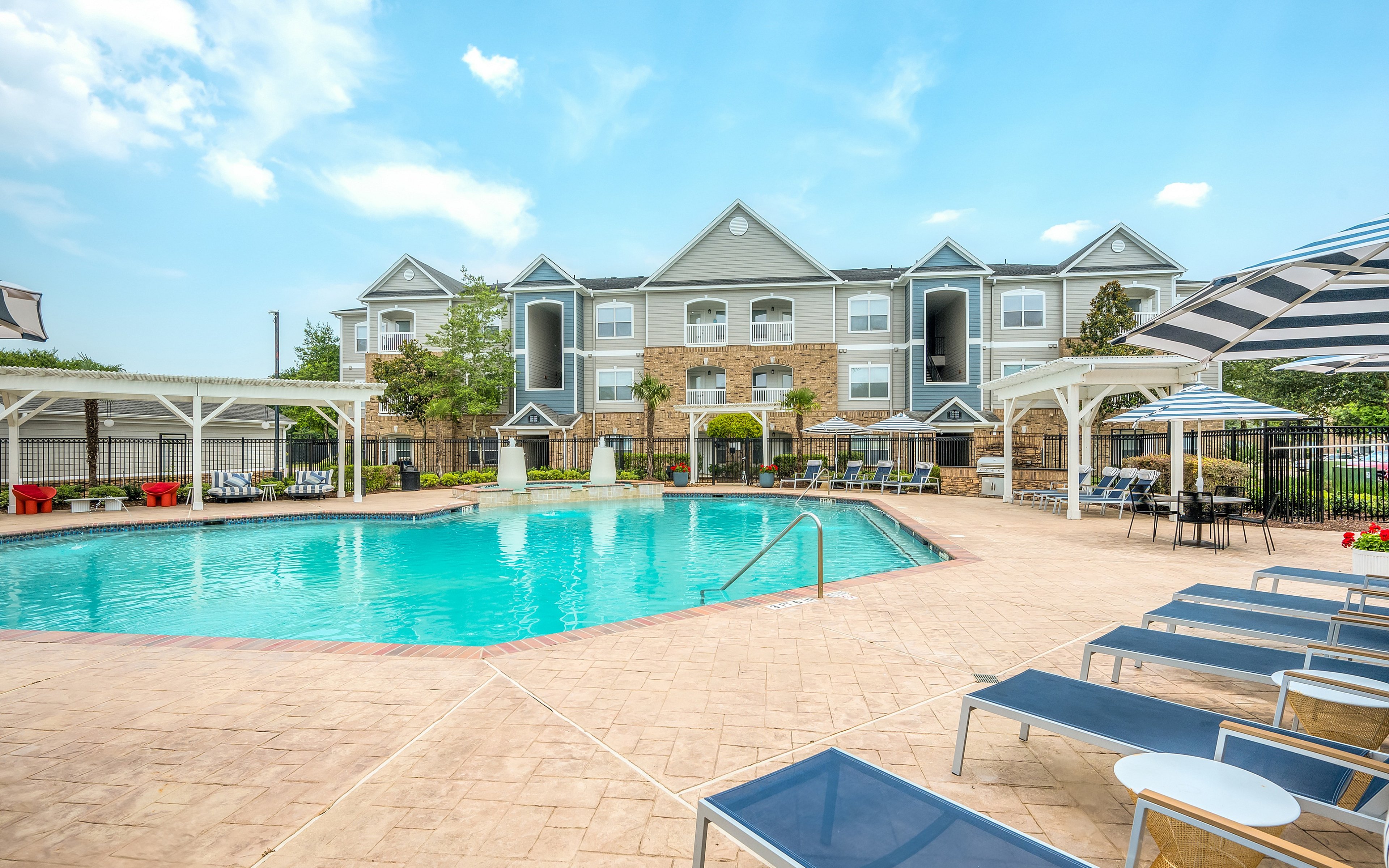 Luxury apartment complex with a sparkling pool surrounded by comfortable lounge chairs, umbrellas, and lush greenery under a clear blue sky.