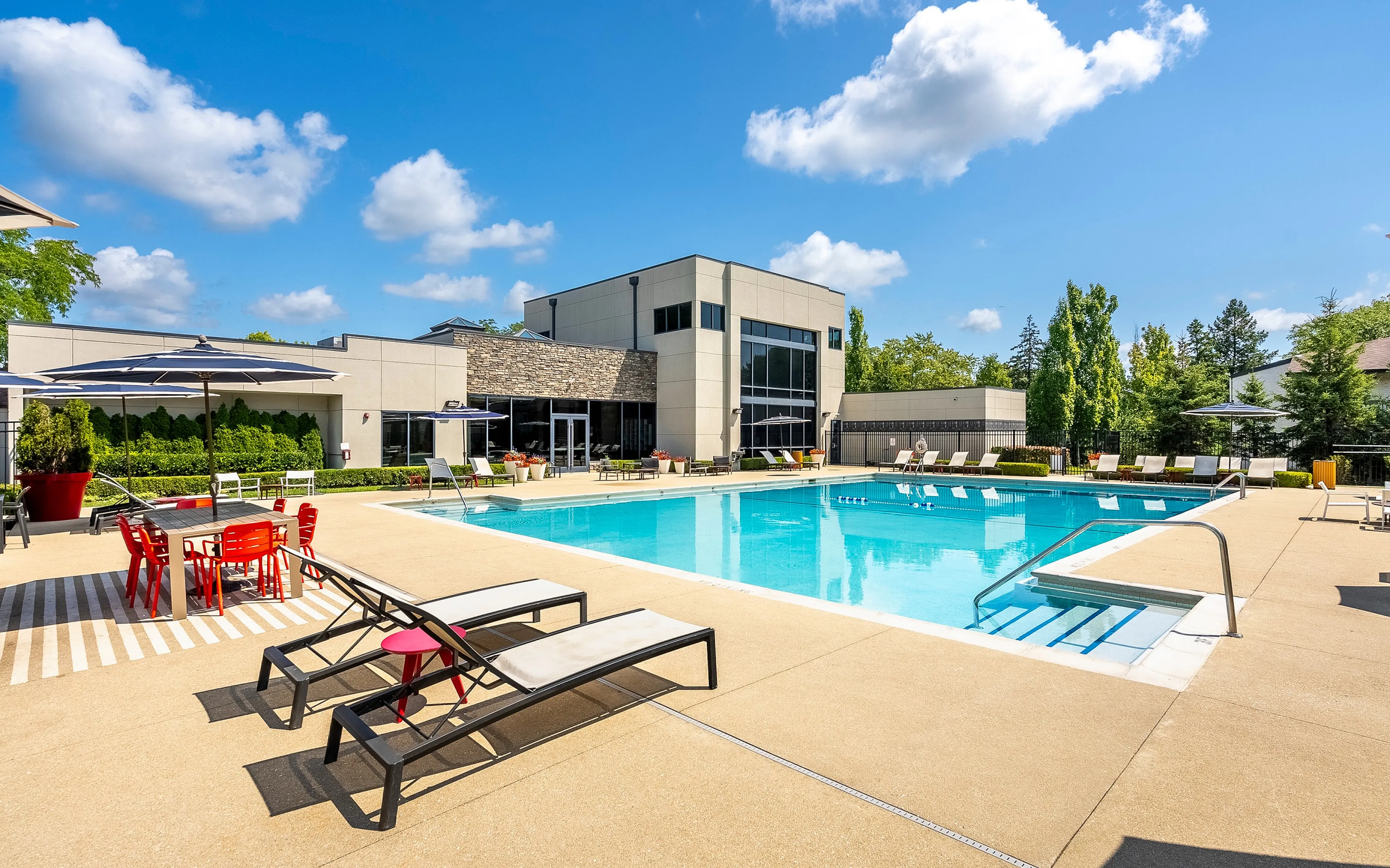 Modern apartment complex featuring a luxurious outdoor pool area with stylish lounge chairs and vibrant red patio furniture under large umbrellas, surrounded by lush greenery and contemporary architecture.
