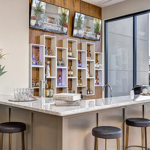 Elegant home bar with a sleek marble countertop, modern stools, and open shelving for drinks, bathed in natural light from large windows.