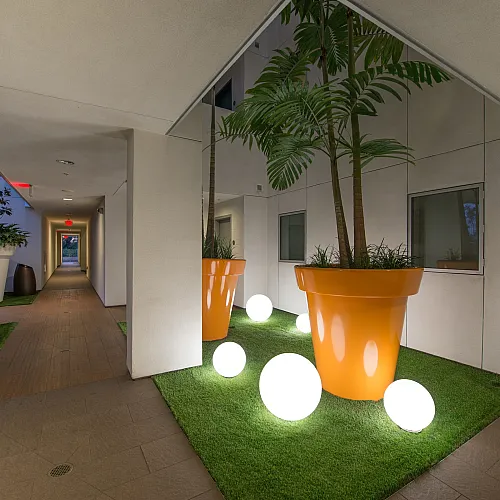 Modern apartment corridor with vibrant orange planters and glowing spherical lights on lush green grass, creating a stylish and inviting atmosphere.