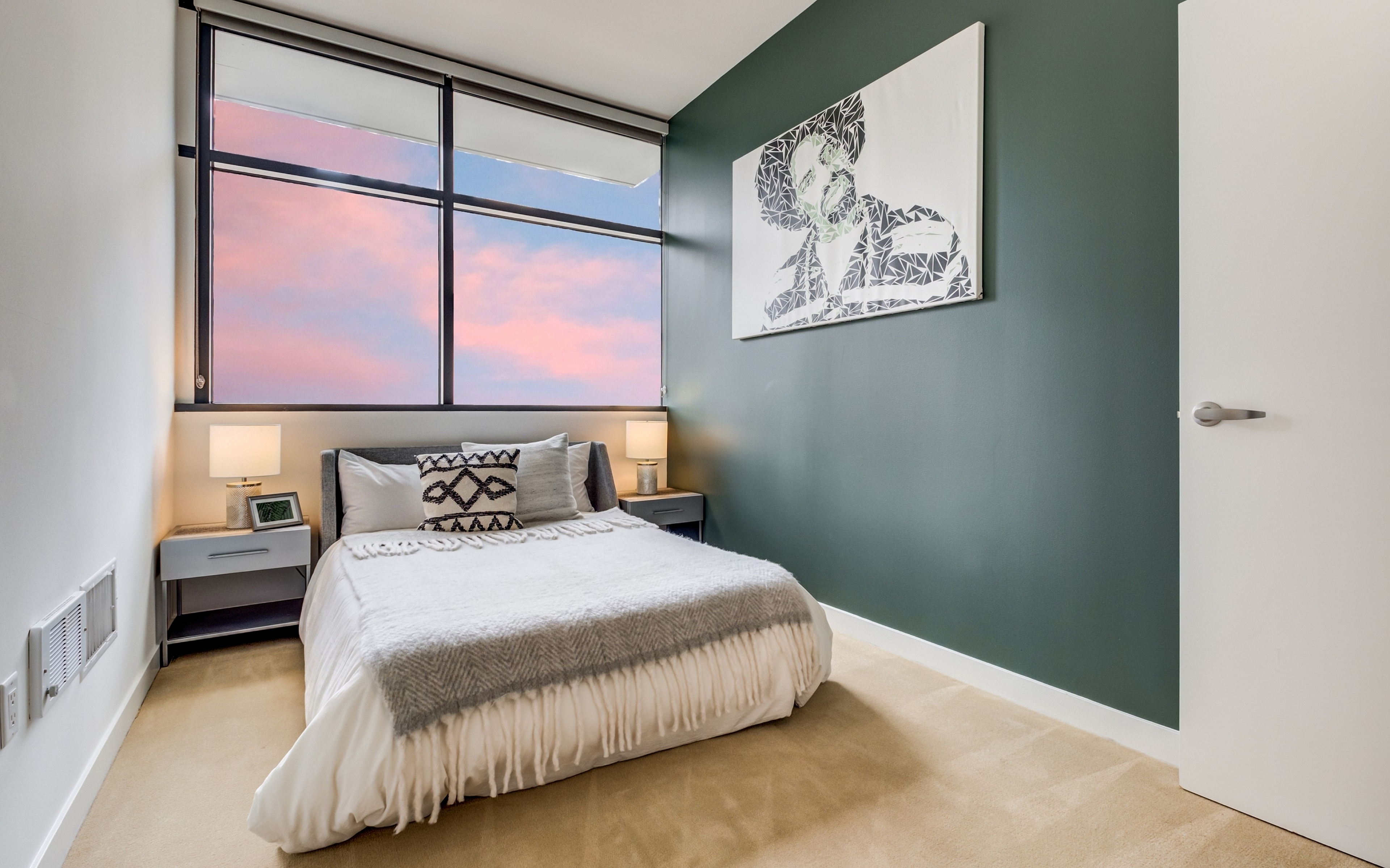 Chic bedroom with a cozy bed, elegant green accent wall, and large window showcasing a stunning sunset view.