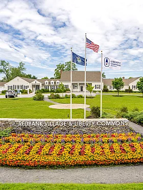Inviting entrance to Redbank Village & Liberty Commons, featuring vibrant flower beds, lush green lawns, and charming architecture under a bright blue sky.
