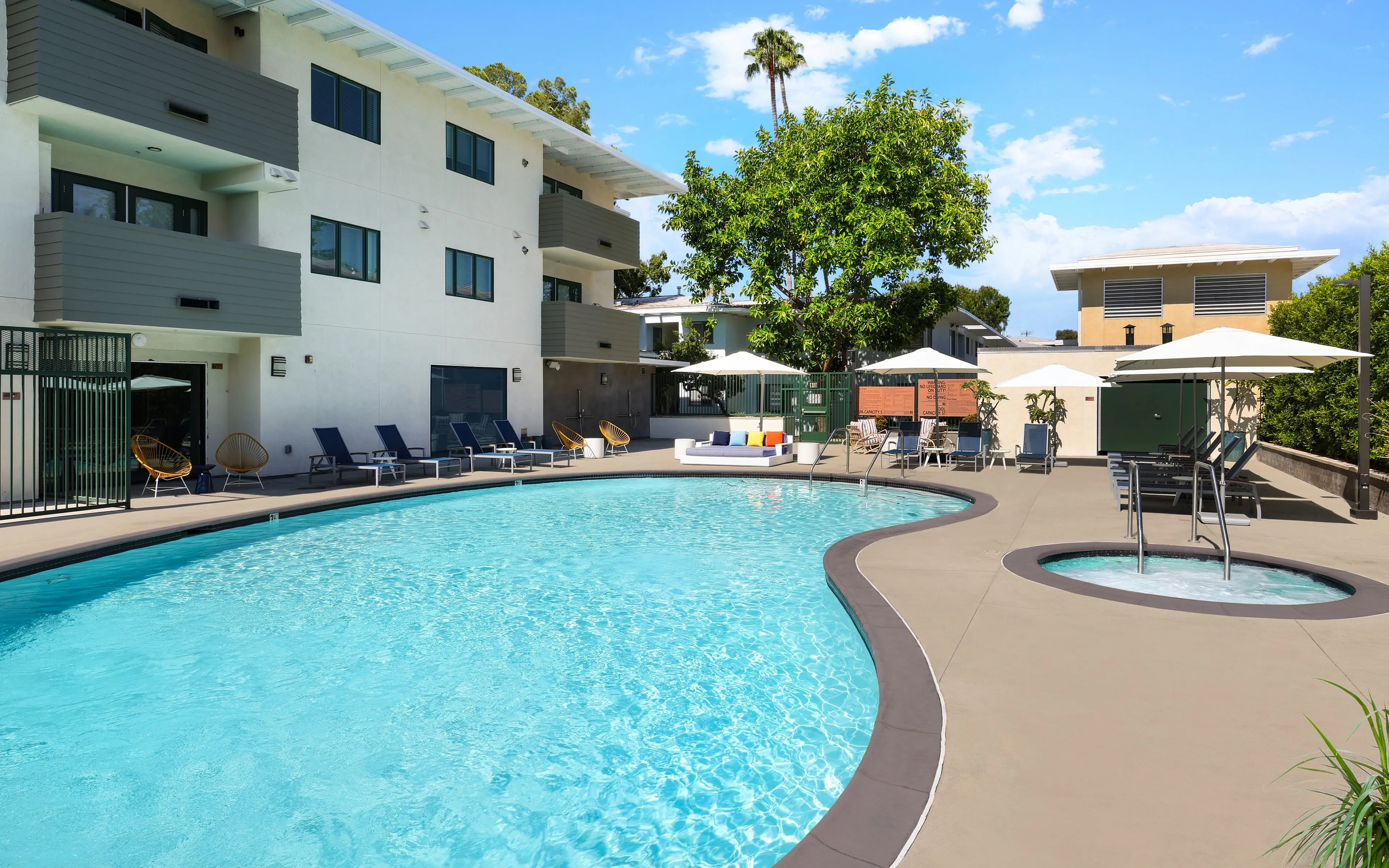 Bright and inviting apartment pool area featuring a sparkling blue swimming pool, relaxing sun loungers, and lush greenery.