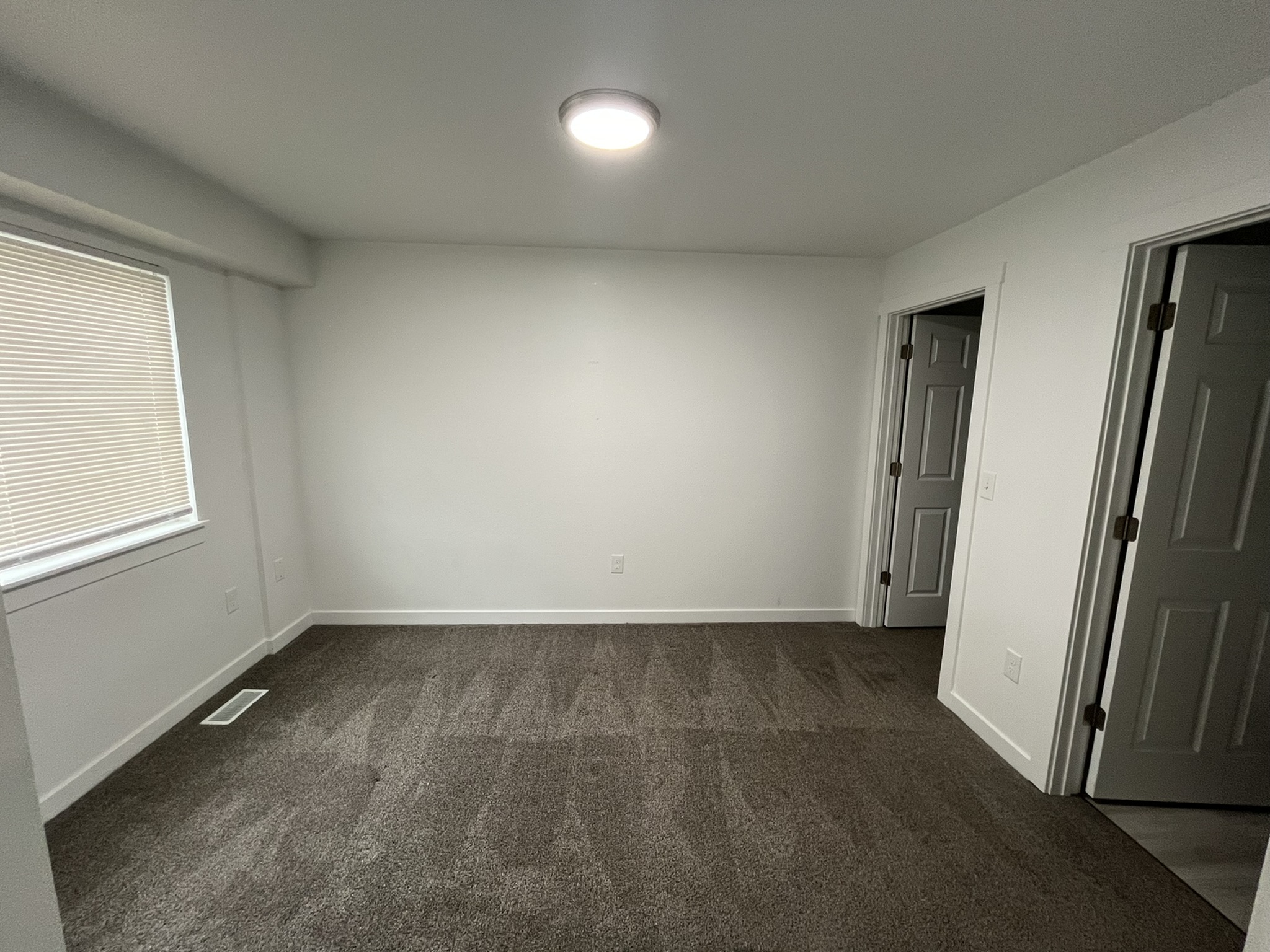 Bright and spacious room with clean carpet, fresh white walls, and ample natural light from a large window, featuring two doors for versatile access.