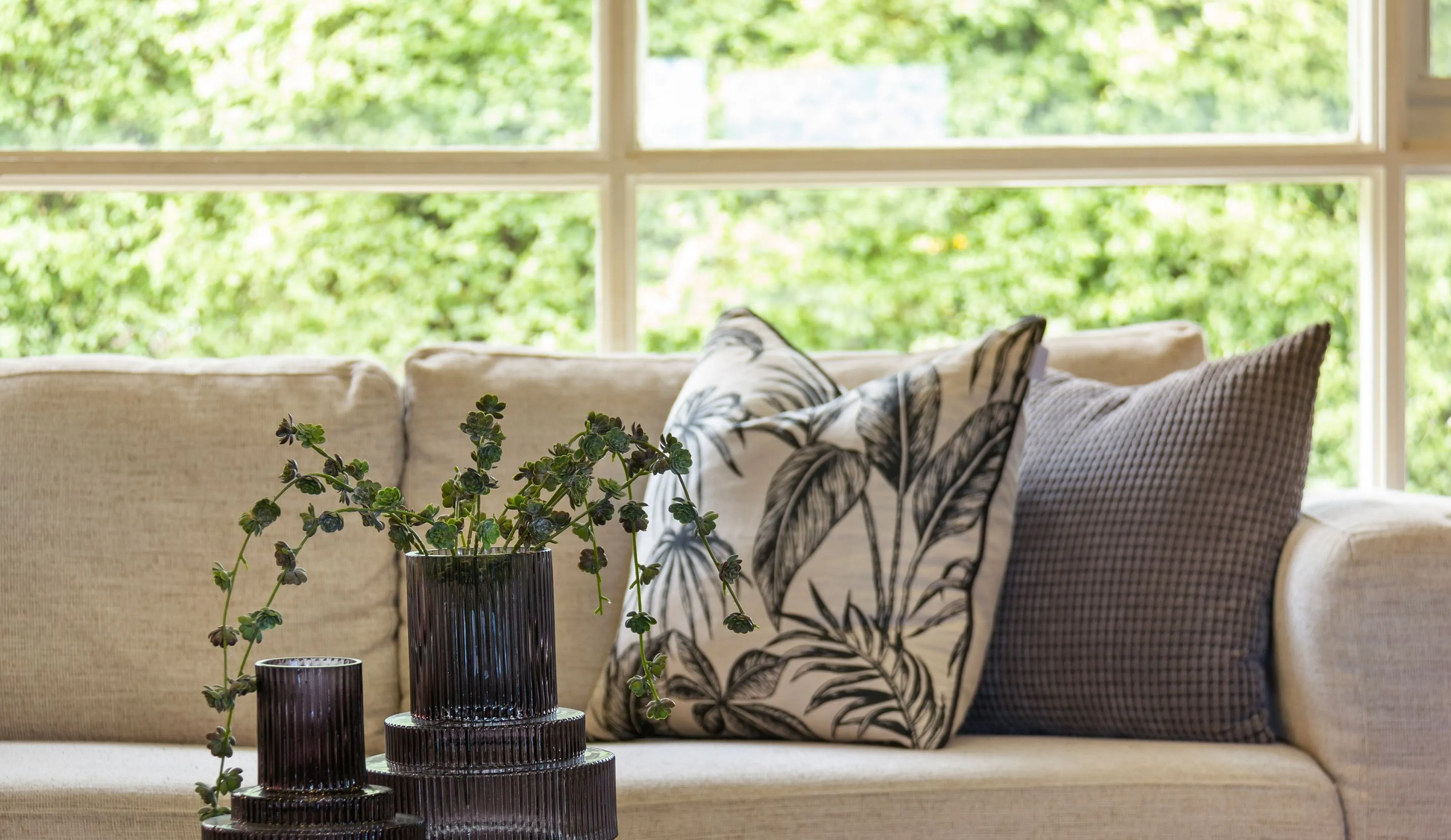 Chic living room with cozy beige sofa, stylish monochrome pillows, and elegant glass vases, framed by a large window with lush green views.