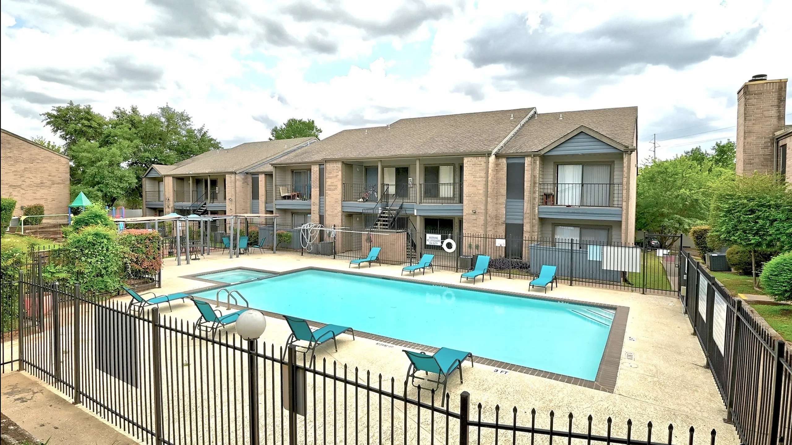Inviting apartment community featuring a sparkling swimming pool surrounded by sun loungers, set against stylish two-story buildings and lush greenery.