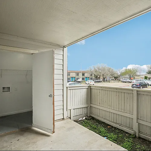 Charming apartment porch with light fencing, offering a spacious view of the parking area under a vibrant blue sky.
