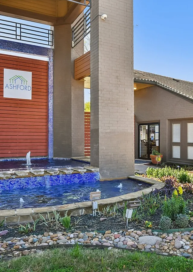 Elegant apartment entrance with a stunning water feature, surrounded by vibrant landscaped gardens and sleek modern architecture.
