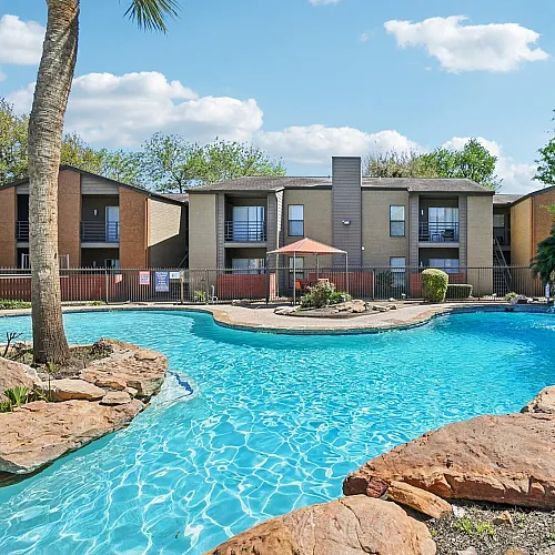 Inviting apartment complex with a stunning pool and waterfall feature surrounded by lush greenery and natural rock formations.