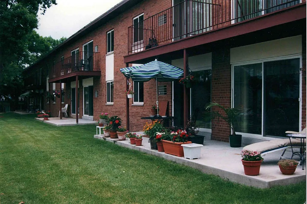 Charming brick apartment exterior with lush green lawn, vibrant flower pots, and cozy patio seating for a serene outdoor retreat.