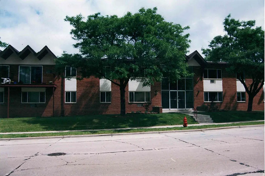 Charming suburban brick apartment building with lush green trees and a manicured lawn.