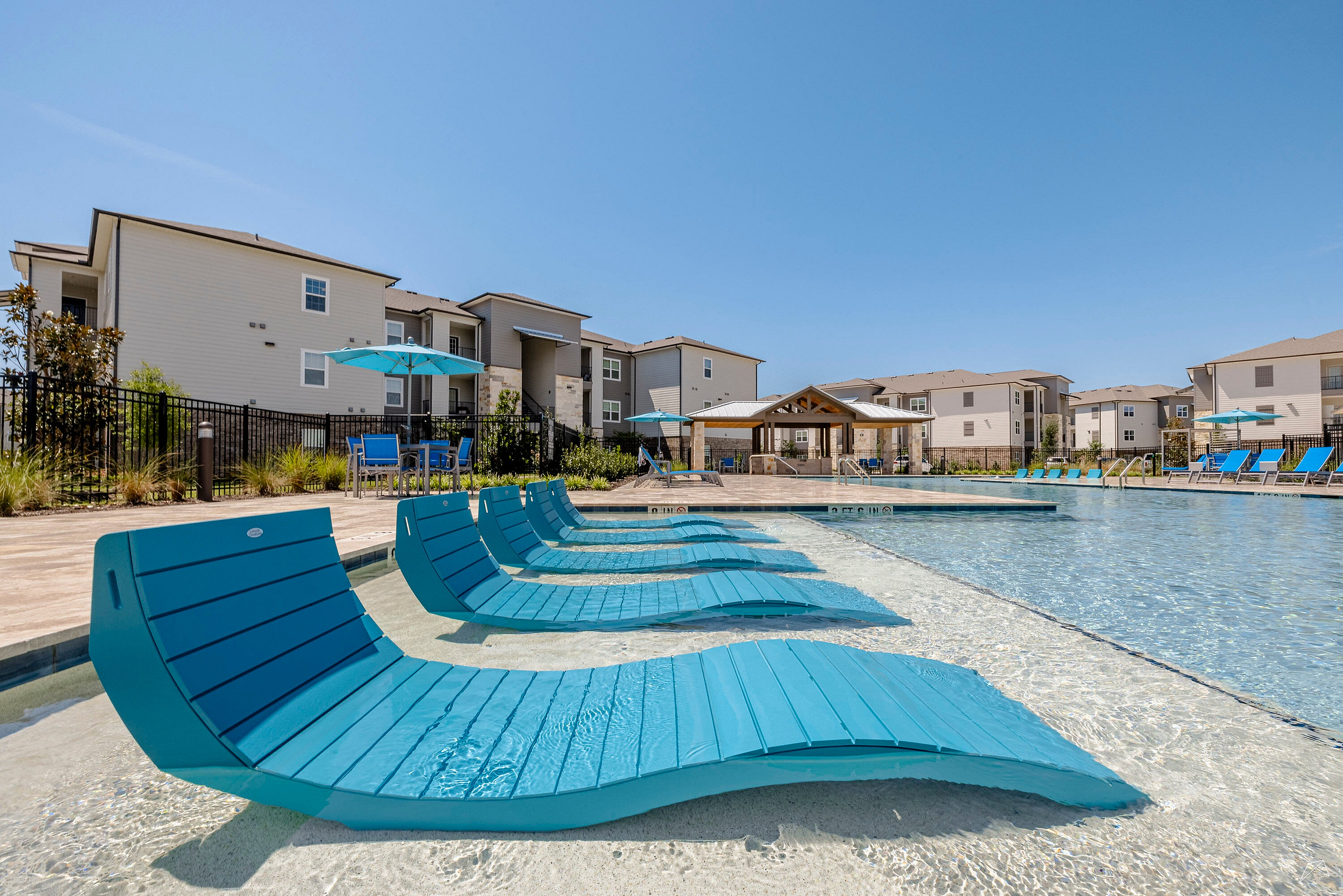 Modern apartment complex featuring a luxurious outdoor pool with stylish blue loungers and shade umbrellas, offering a perfect blend of relaxation and comfort.
