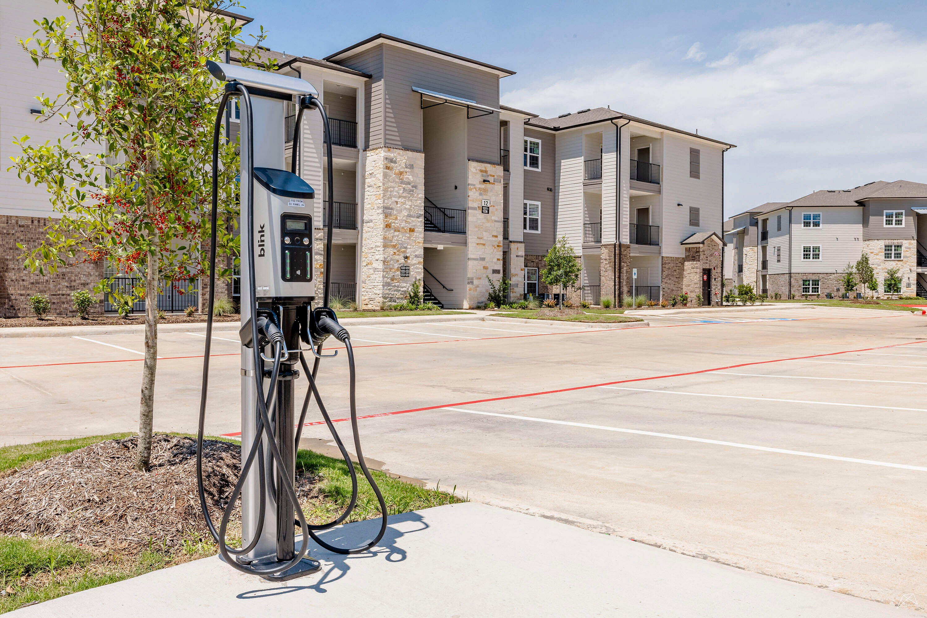 Modern apartment complex with elegant stone facades and outdoor EV charging station, surrounded by well-maintained landscaping and spacious parking lot.