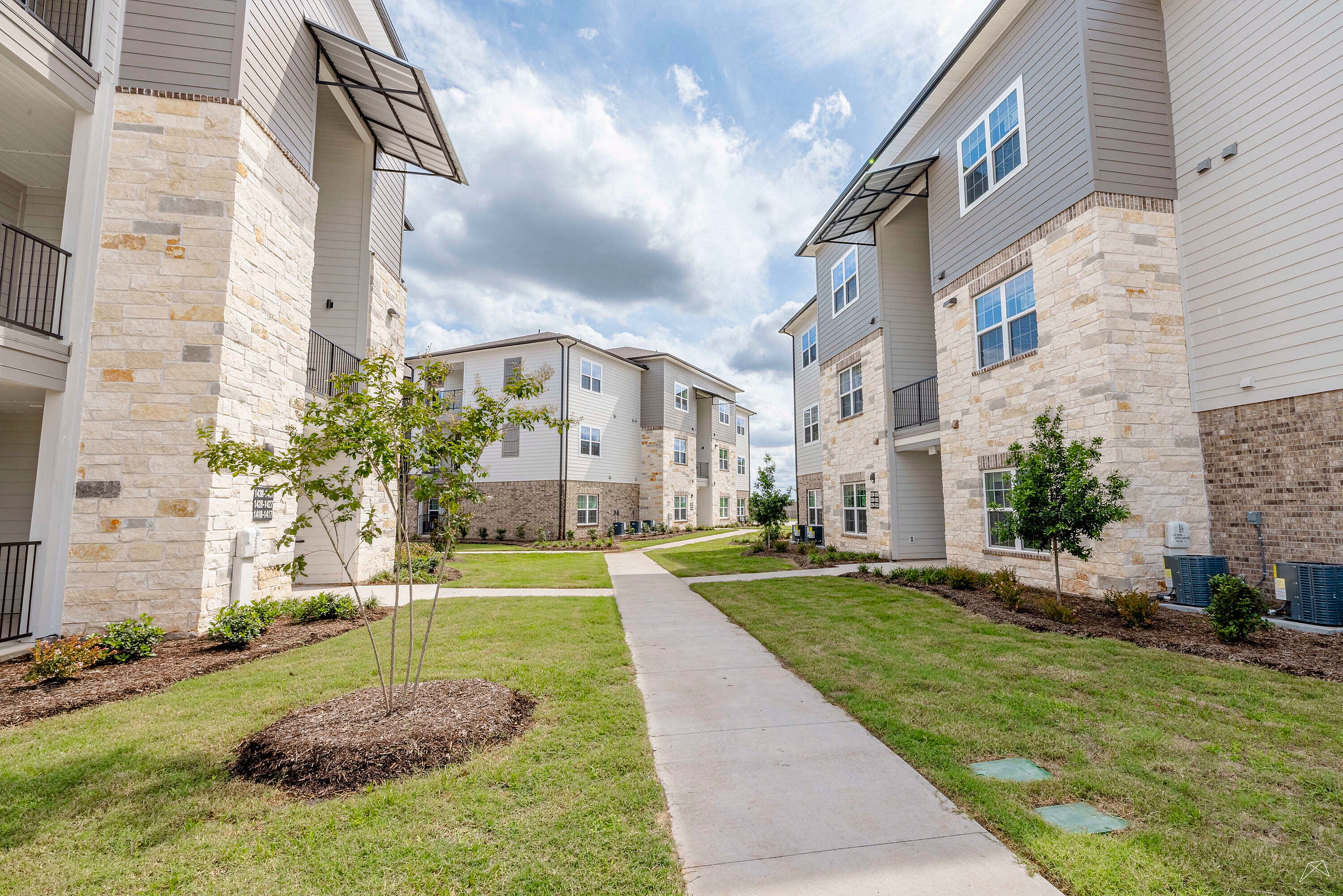 Beautifully landscaped apartment complex with modern stone and panel exterior, clear pathways, and lush greenery under a bright sky.