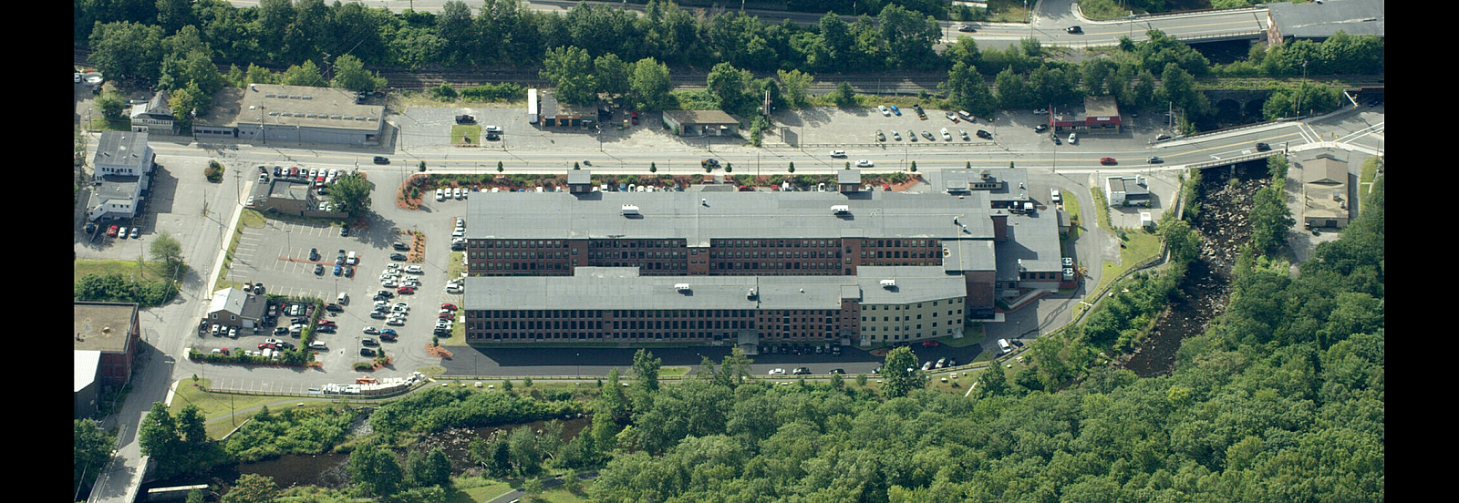Aerial view of a large apartment complex surrounded by lush greenery and a serene river, offering ample parking and scenic views.