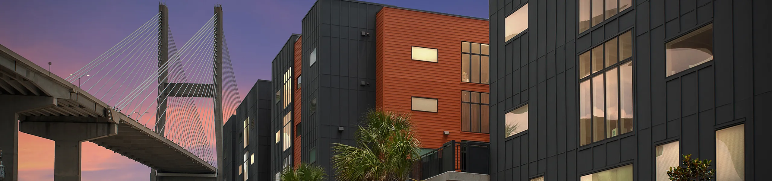 Modern urban apartment building with sleek black and orange facades, next to an impressive cable-stayed bridge at sunset.