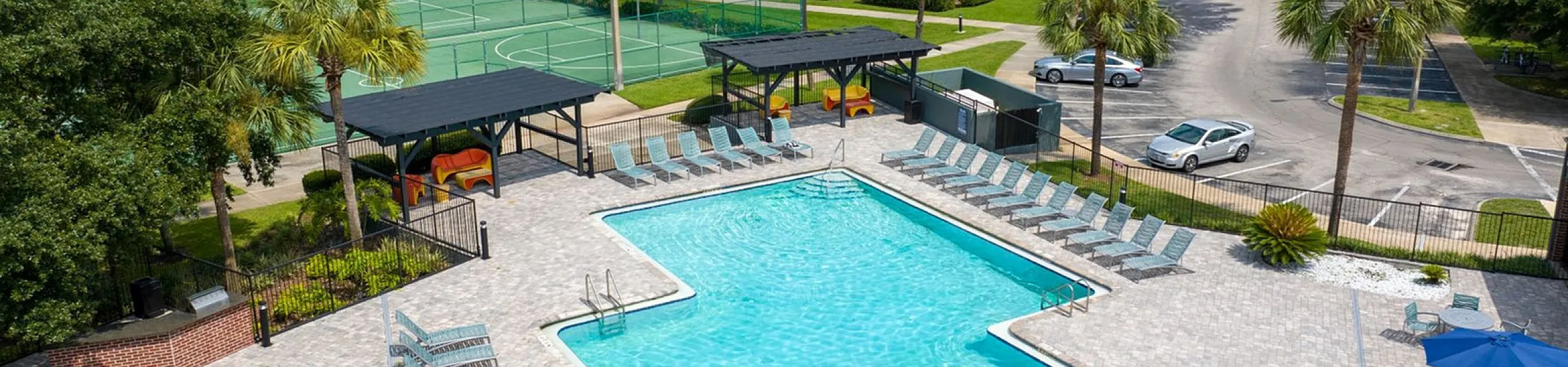 Inviting apartment complex featuring a sparkling swimming pool, sun loungers, shaded seating areas, and a tennis court surrounded by lush greenery.
