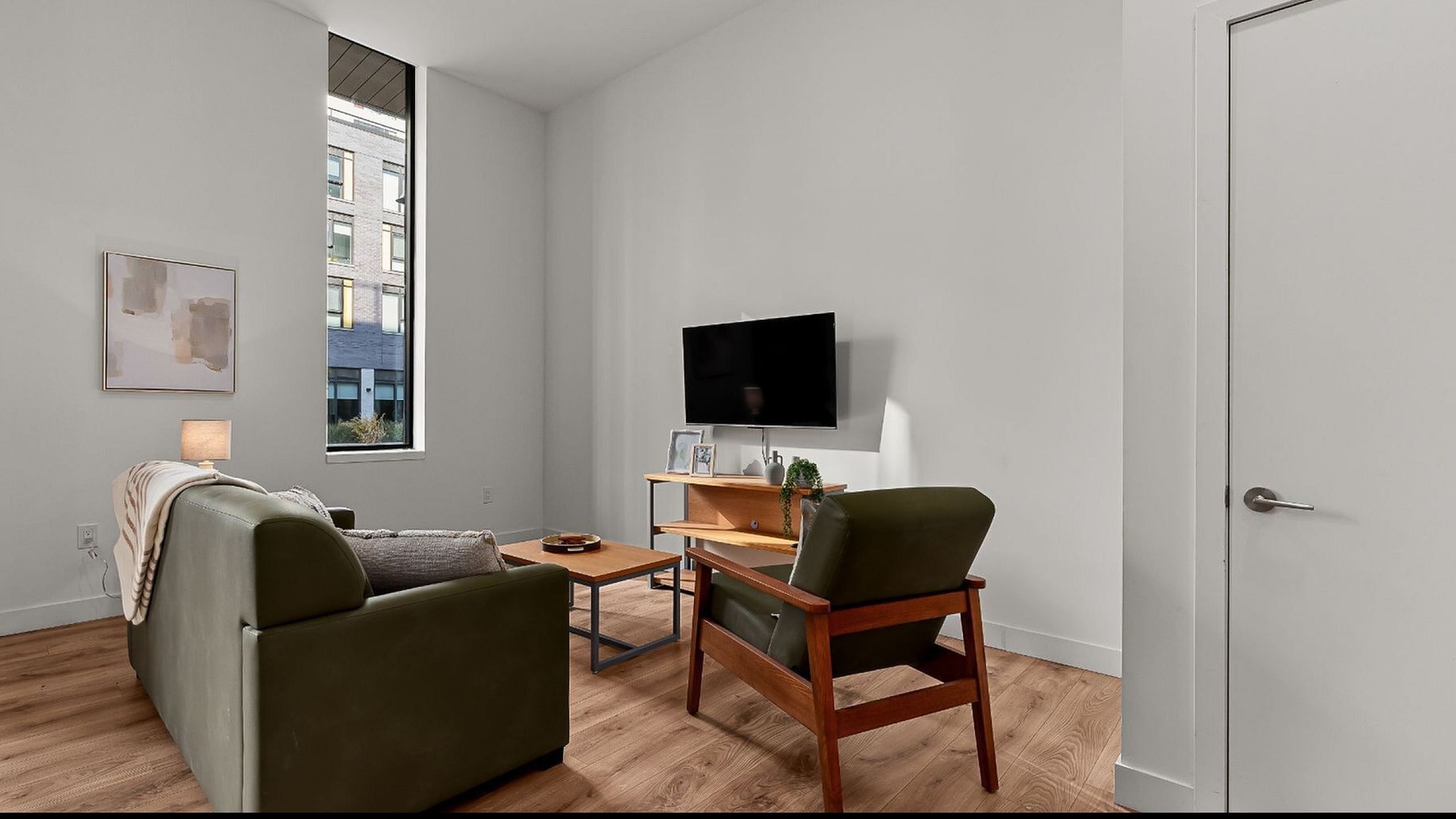 Bright and modern living room with stylish furniture, a wall-mounted TV, and large window offering city views.