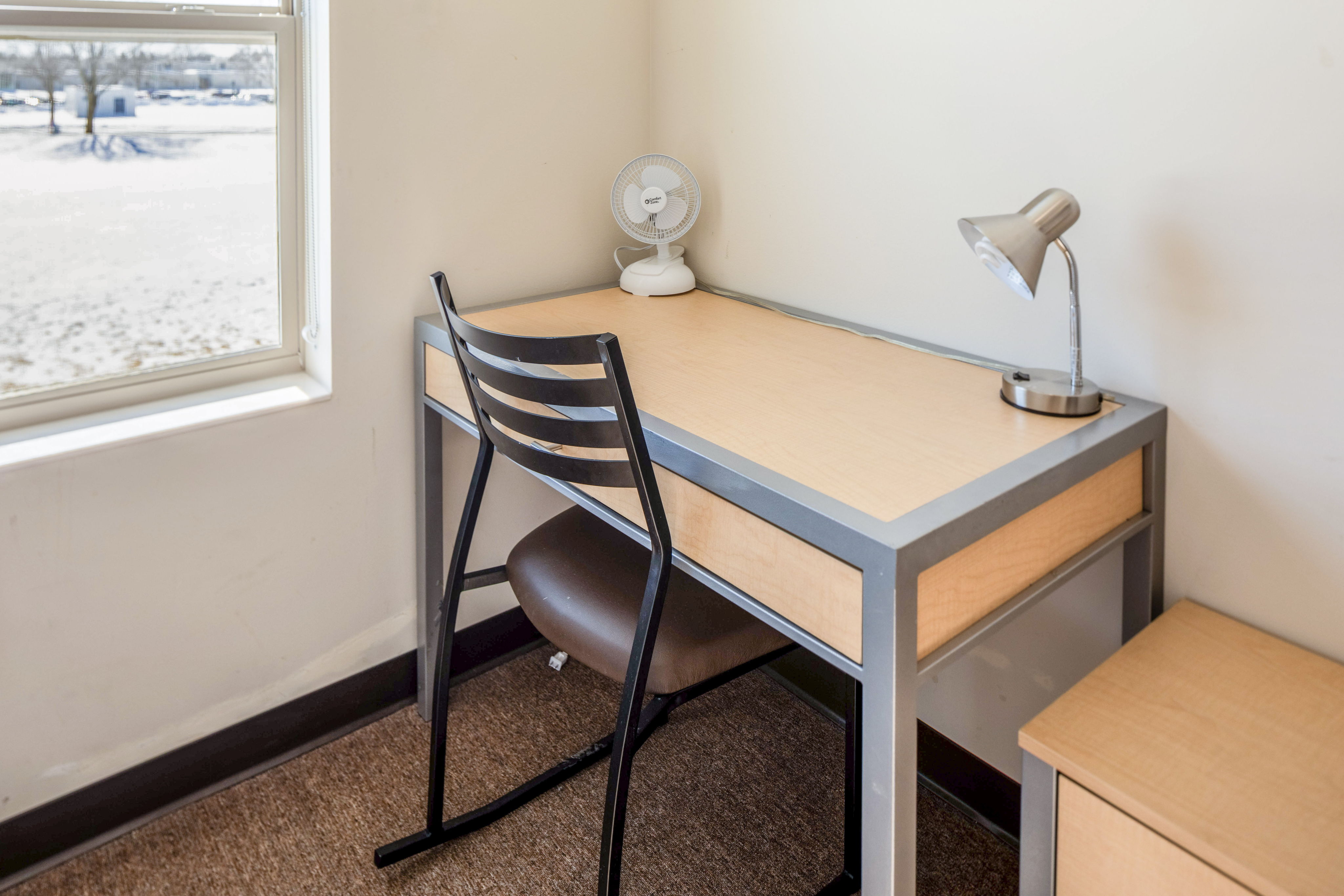 Bright corner with a wooden desk, modern chair, lamp, and small fan near a sunny window.