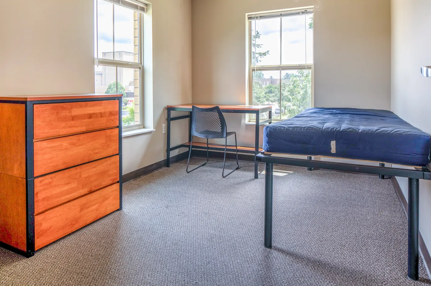 Bright and inviting bedroom with a modern desk, comfortable bed, and stylish dresser, bathed in natural light from large windows overlooking a lush view.