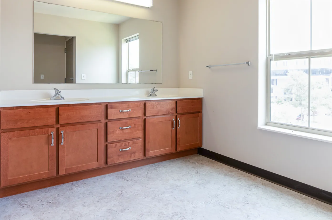 Bright, spacious bathroom featuring a double vanity with elegant wooden cabinetry and a large mirror, complete with a sunny window view.