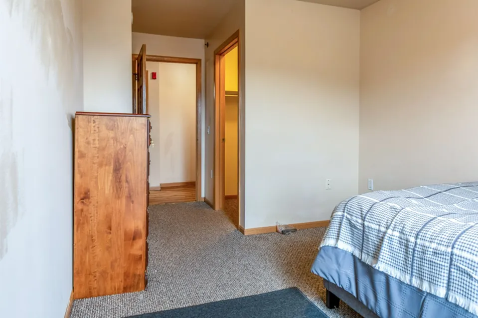 Inviting bedroom with cozy carpet flooring, wooden dresser, and a soft checkered bedspread for a relaxing atmosphere.