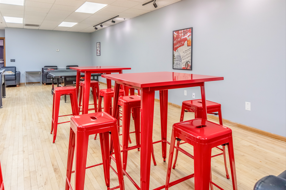 Vibrant communal area with striking red high tables and stools, perfect for social gatherings. Bright, spacious room with hardwood floors and modern decor.