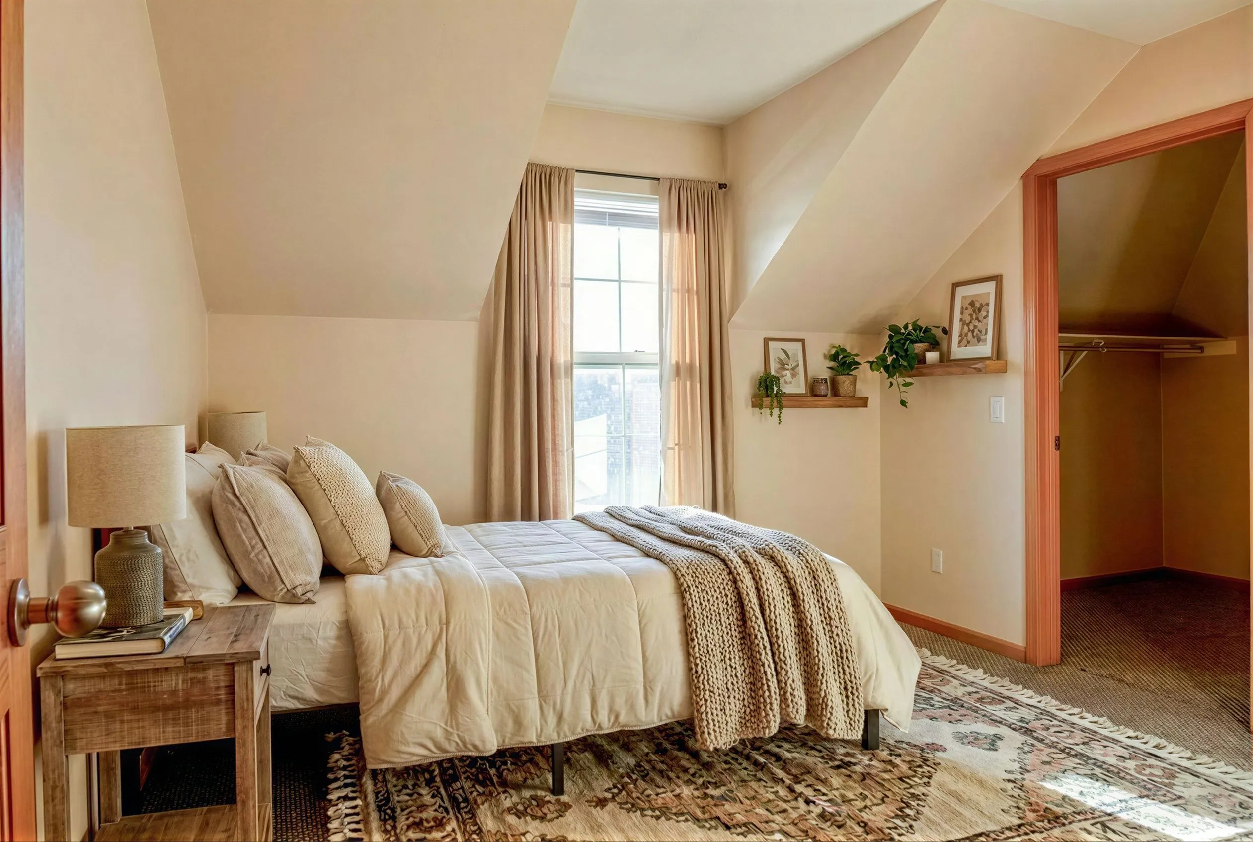 Cozy bedroom retreat featuring elegant beige decor, large window for natural light, and stylishly adorned shelves with plants and art.
