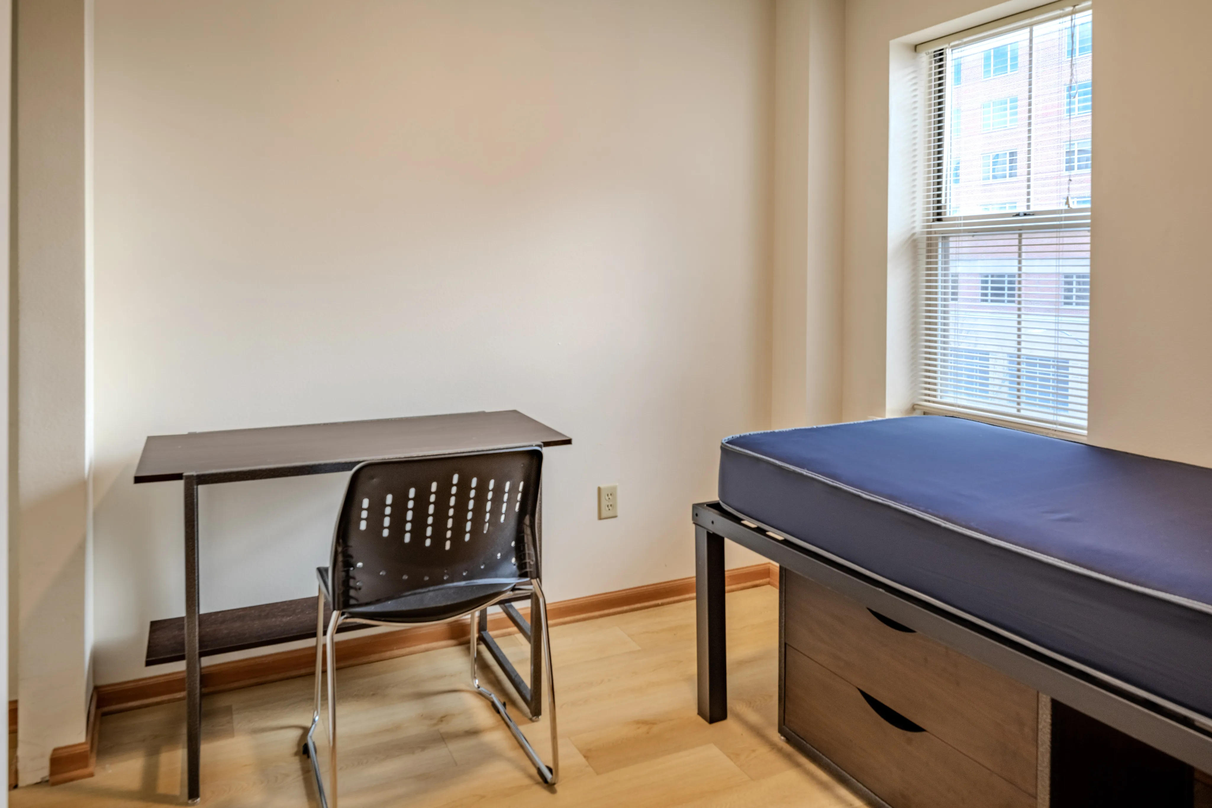 Minimalist bedroom with a single bed, desk, and chair, featuring natural light from a window.