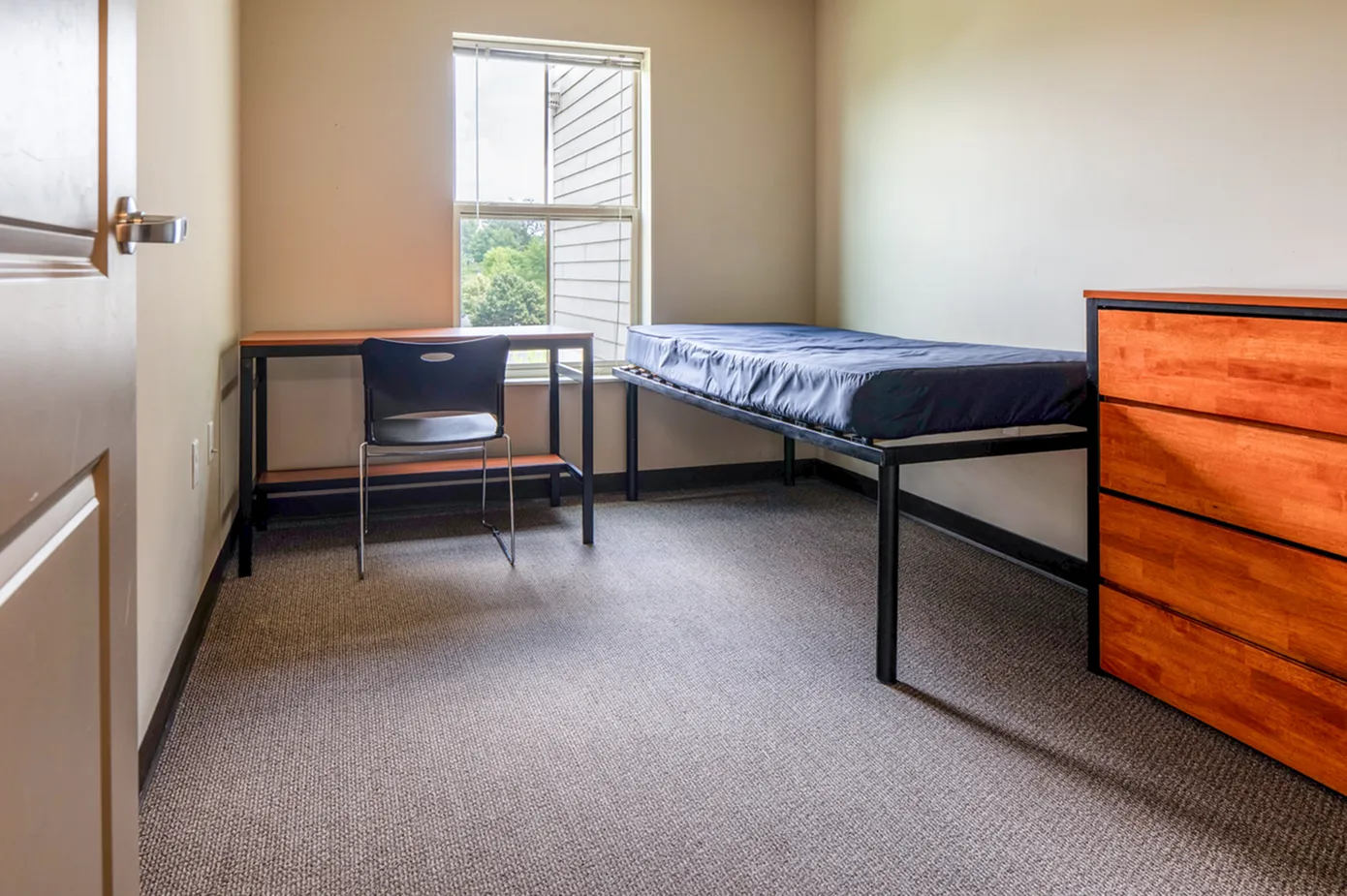 Bright bedroom featuring a single bed with a minimalist design, a functional desk with a chair, and a window offering a view of lush greenery.