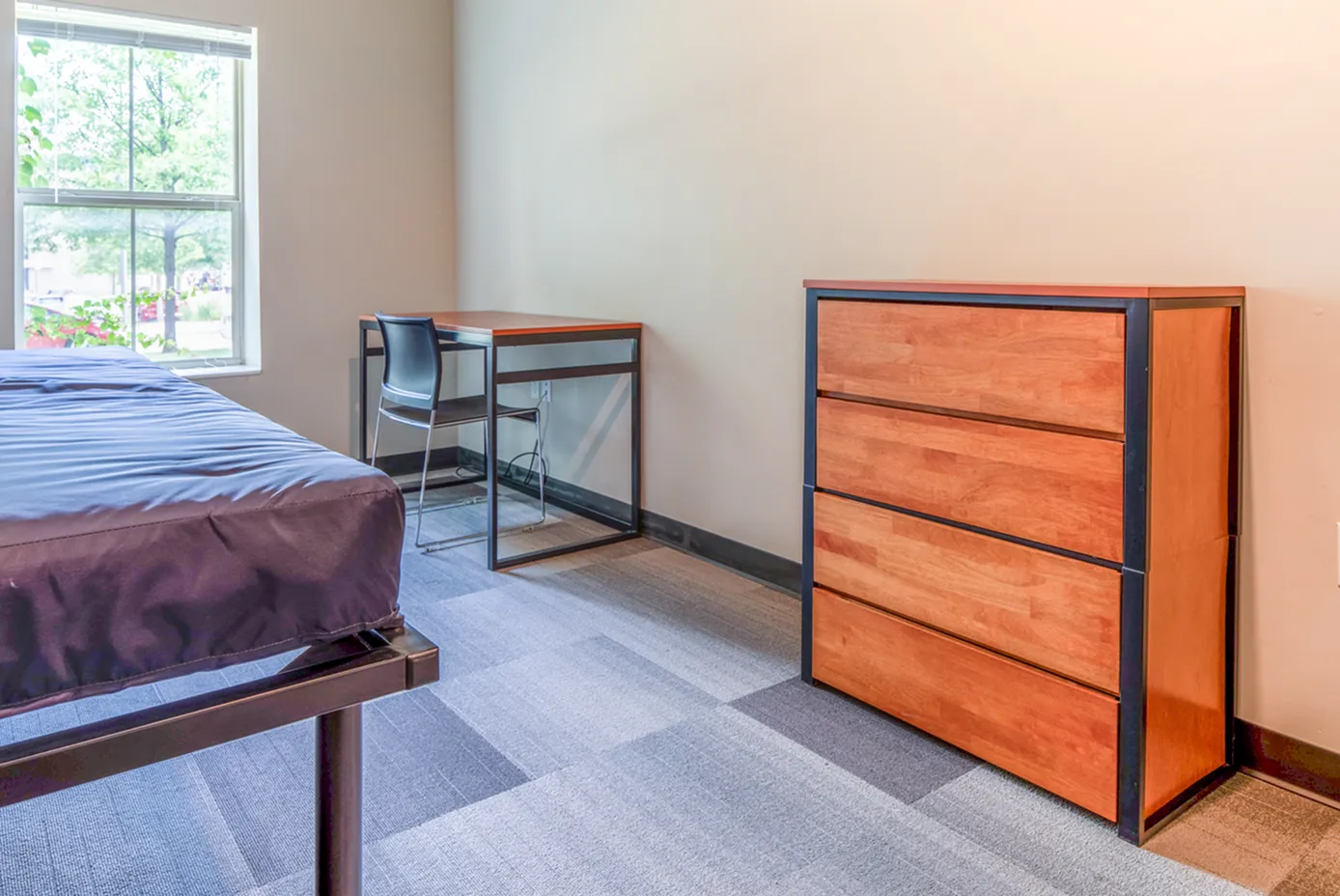 Cozy bedroom with a large window offering natural light, featuring a sleek wooden dresser, a minimalist desk, and a comfortable bed.